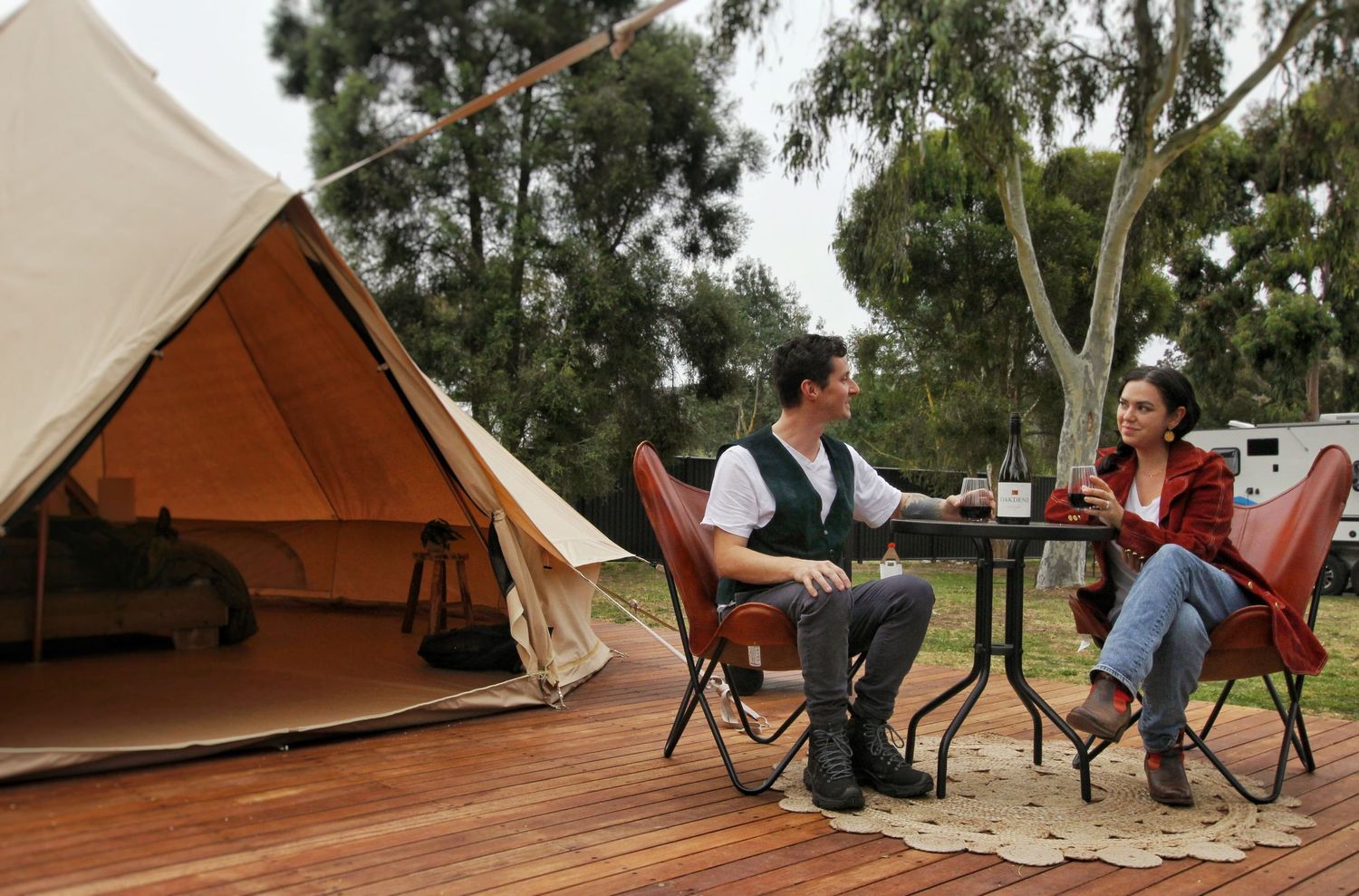 Couple enjoying drinks at a table on a timber deck outside a canvas bell tent surrounded by gum trees.