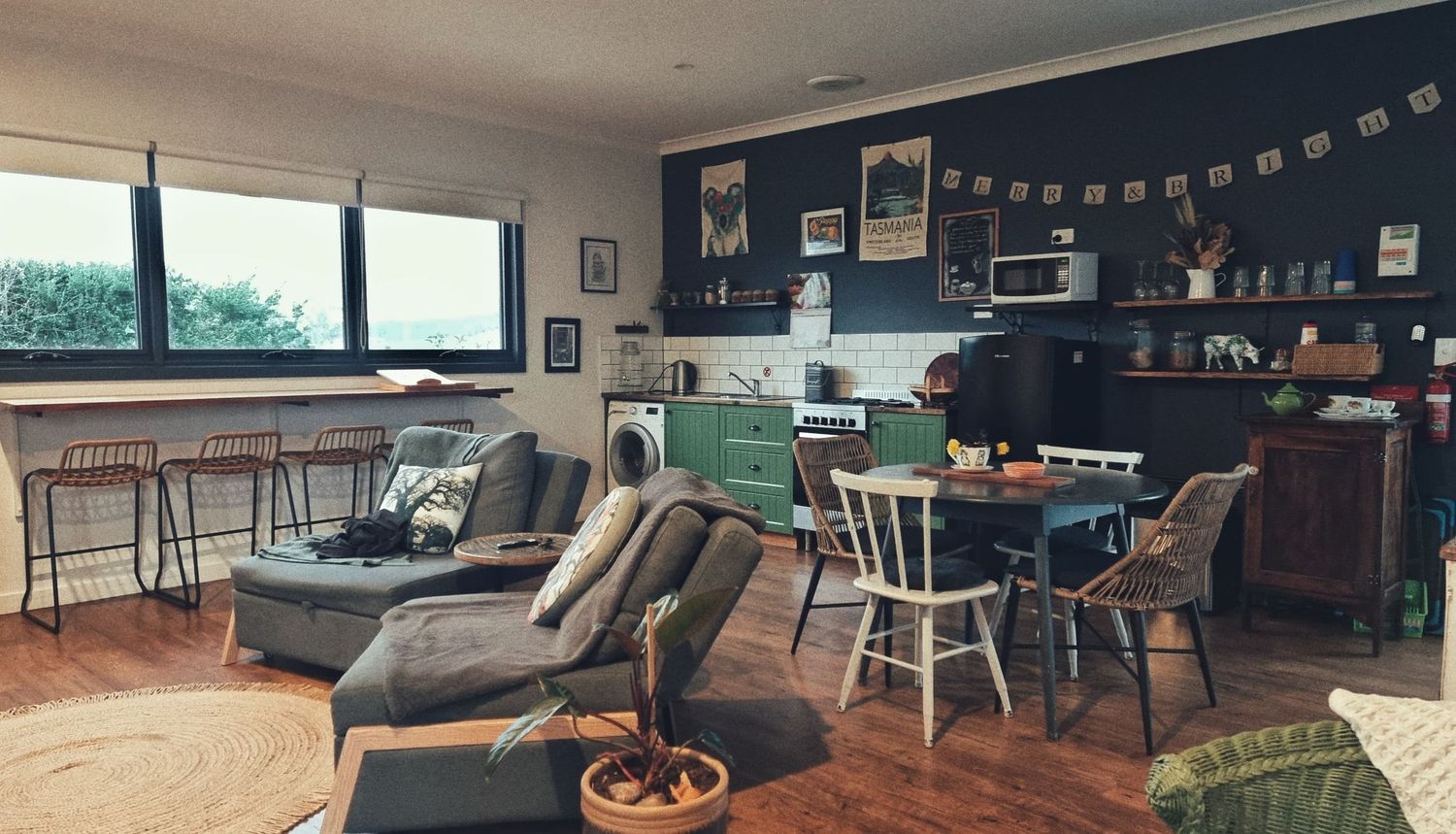 Open-plan cottage interior with a cosy lounge, dark blue feature wall, green kitchen cabinets, dining table and timber floors.