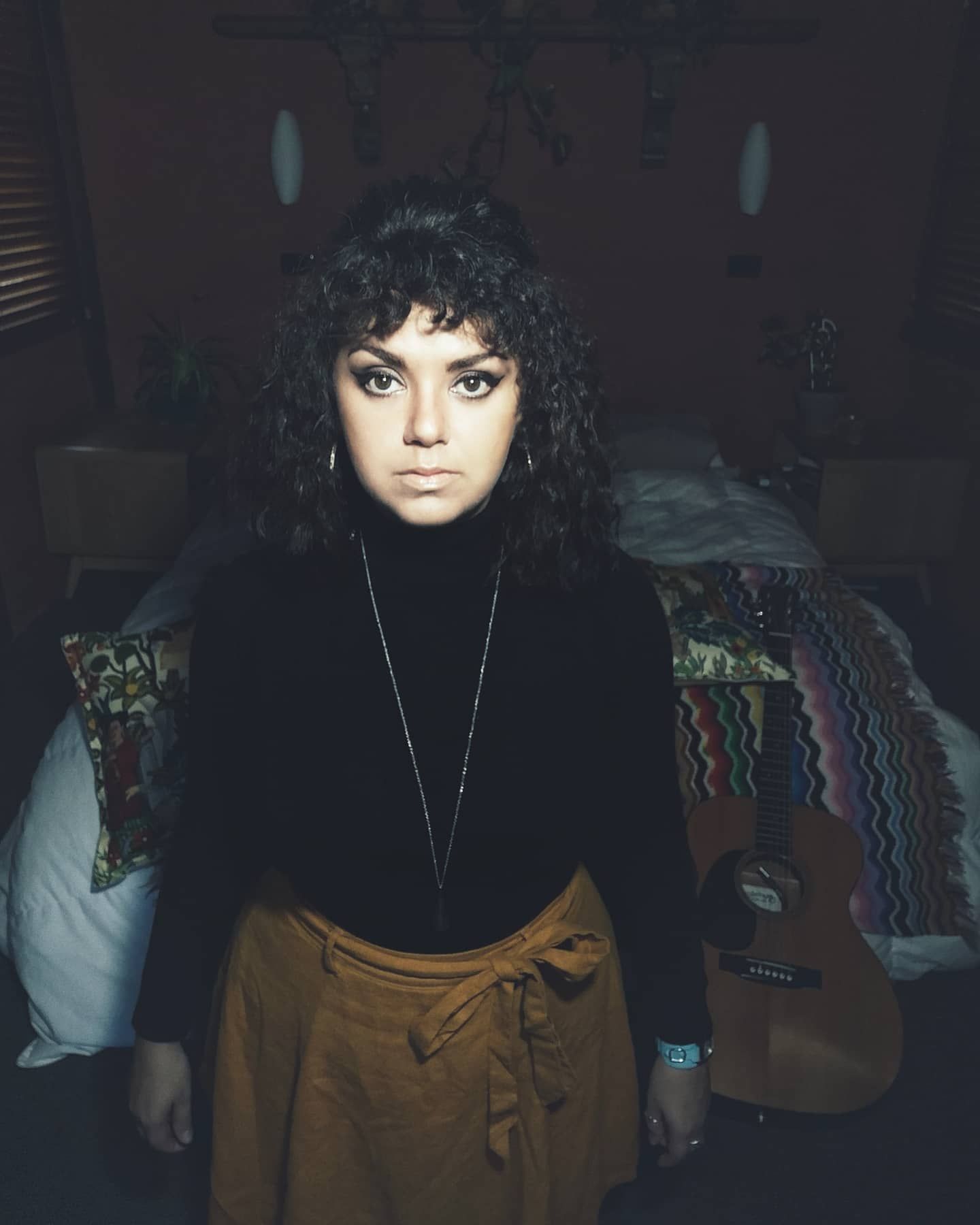 Woman with curly dark hair in a black turtleneck and mustard skirt stands in a dim bedroom with an acoustic guitar behind her.