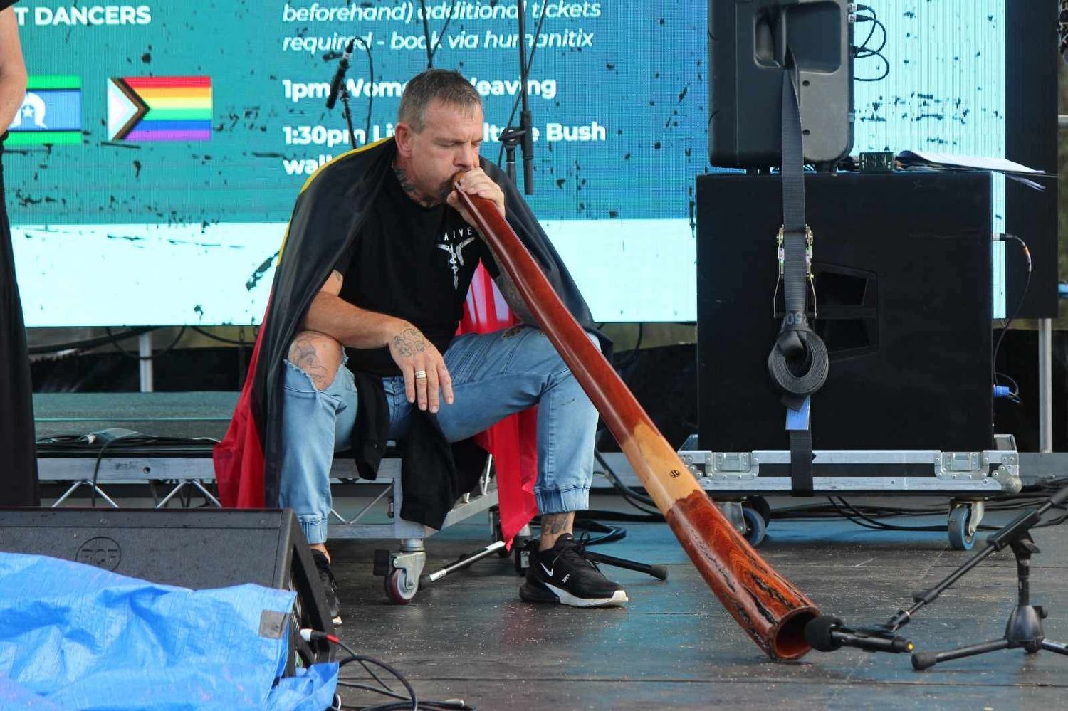 A man in a black vest sits on a festival stage playing a long didgeridoo, with a schedule screen showing dancers behind him.