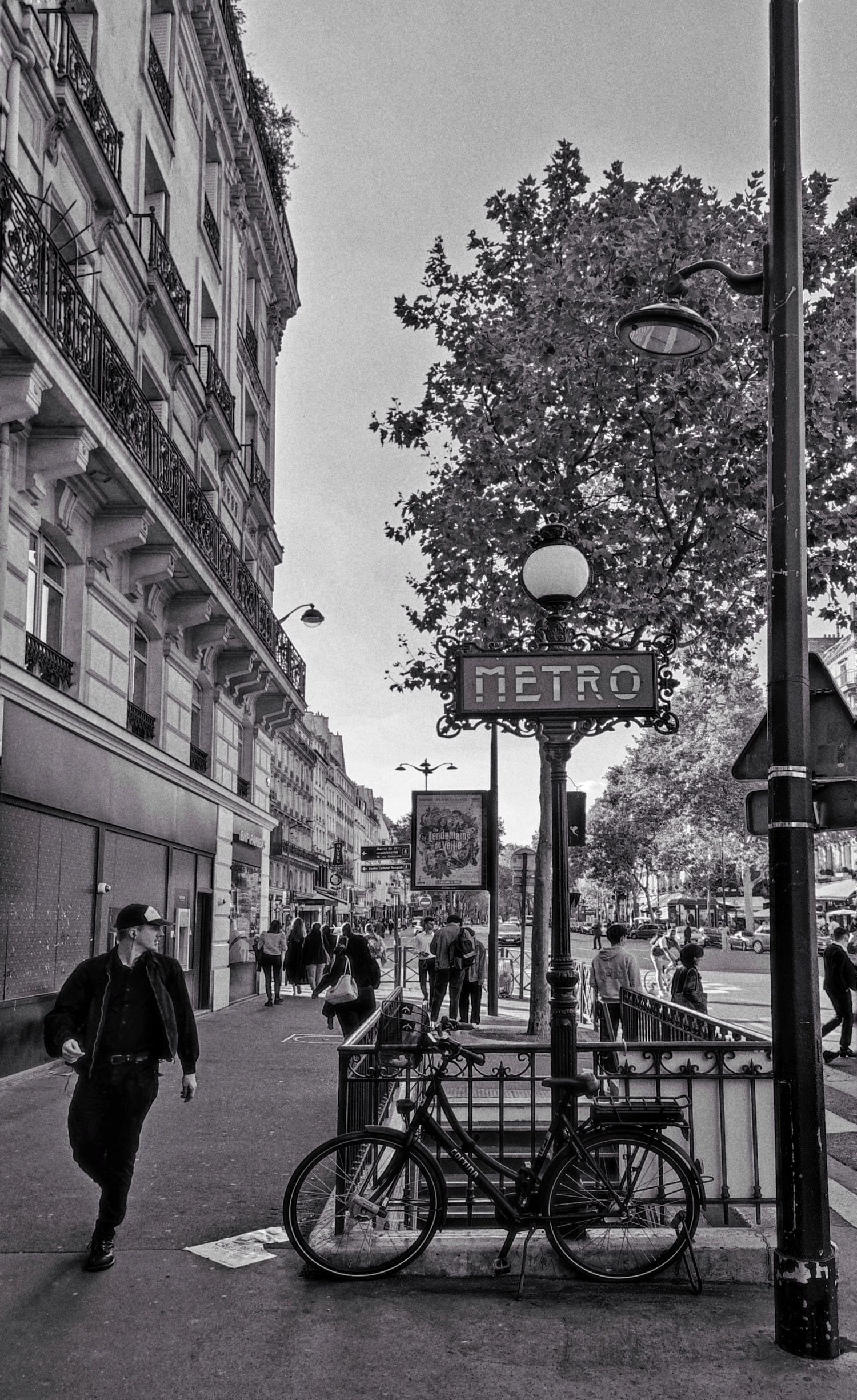 Black and white street scene of a Paris Metro entrance sign, parked bicycle, pedestrians and leafy trees along a Haussmann boulevard.