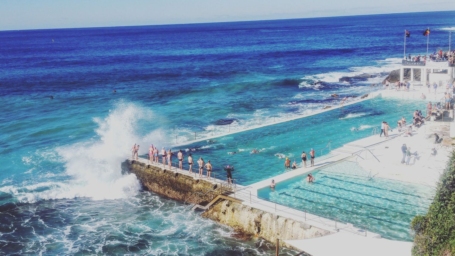 Swimmers and spectators gather at a seaside ocean pool as waves crash against the rocky coastline under a bright blue sky.