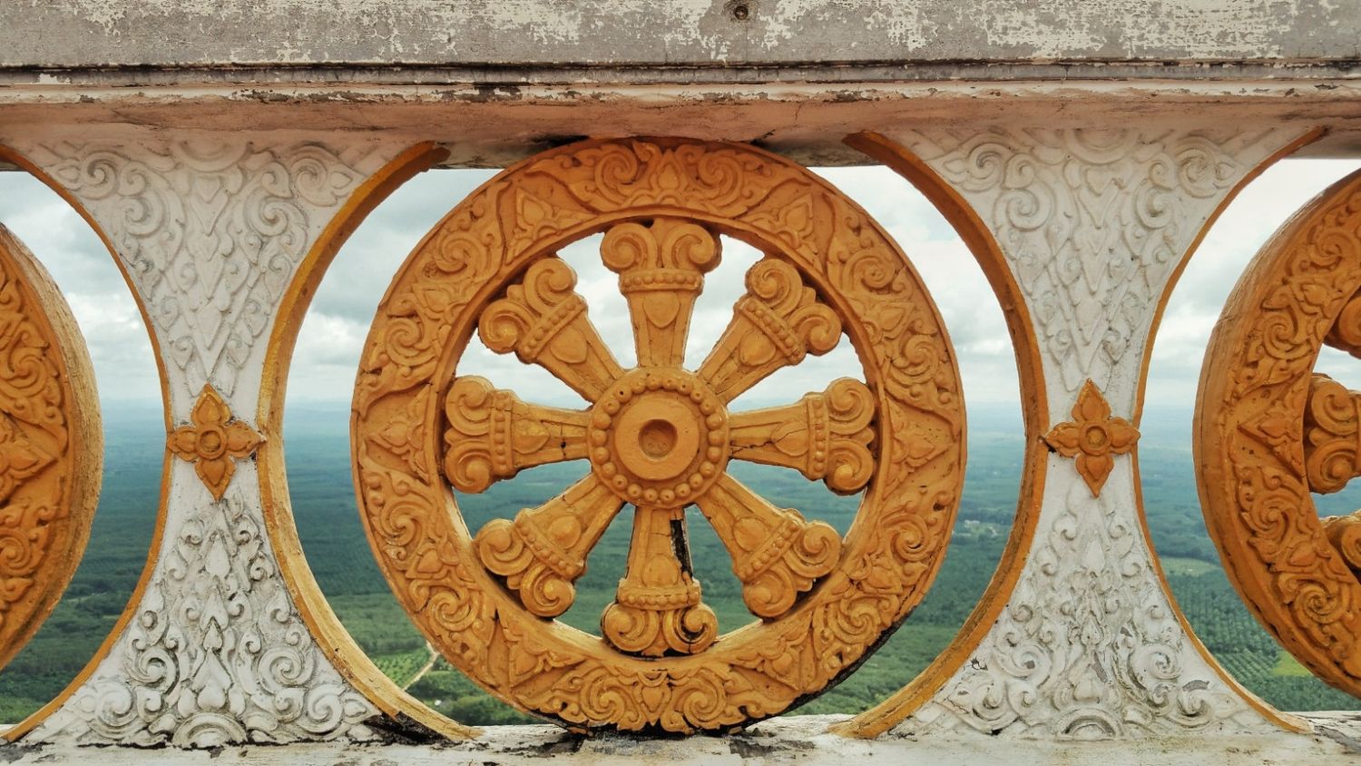 Ornate golden-orange Dharma wheel carving set in a white stone balustrade, with green hills and sky visible behind.