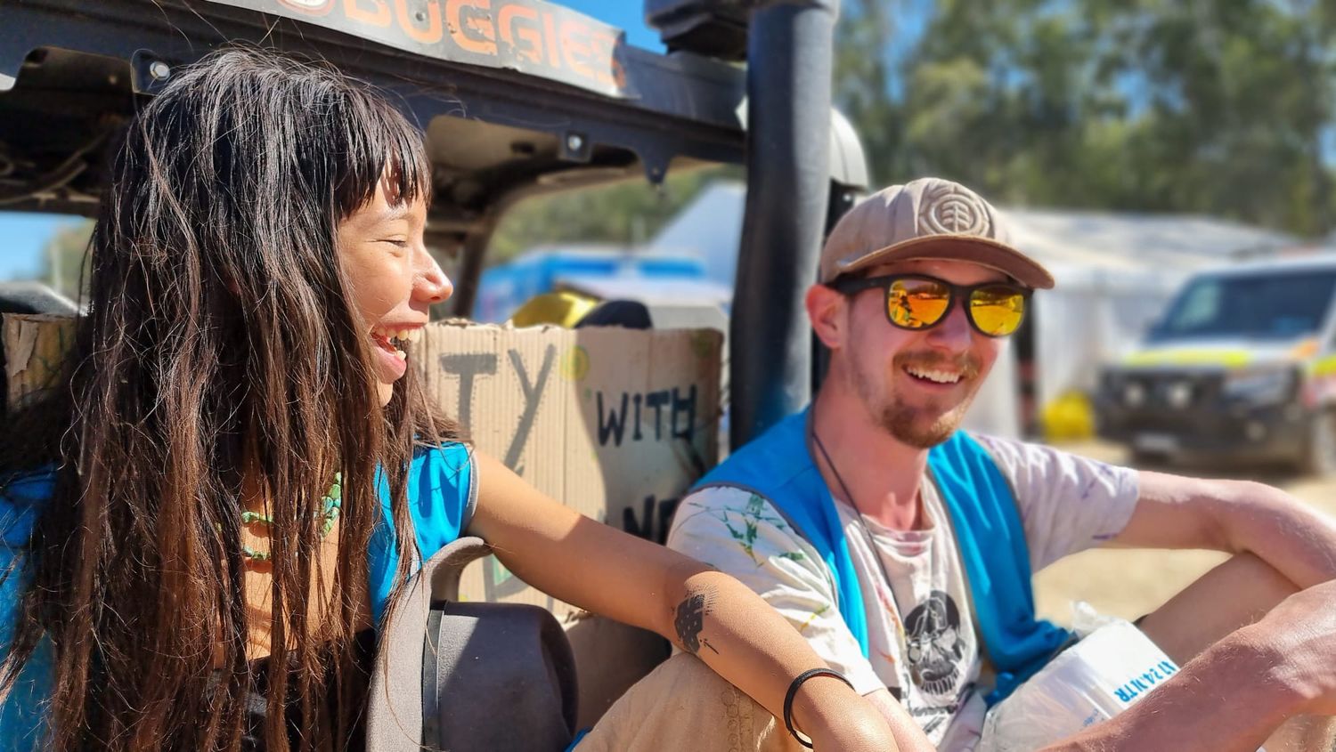 A smiling woman with long dark hair and a man in a cap and orange sunglasses relax together in an open-air vehicle outdoors.