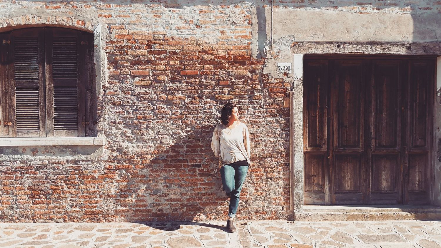 Woman in a white top and jeans leaning against an old red brick wall beside weathered wooden doors.