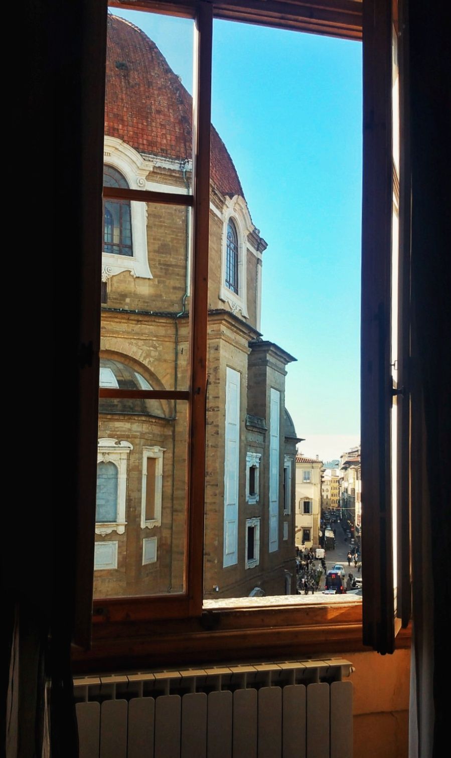 View through an open wooden window of a historic Italian building with a terracotta dome and a sunlit street below under a blue sky.