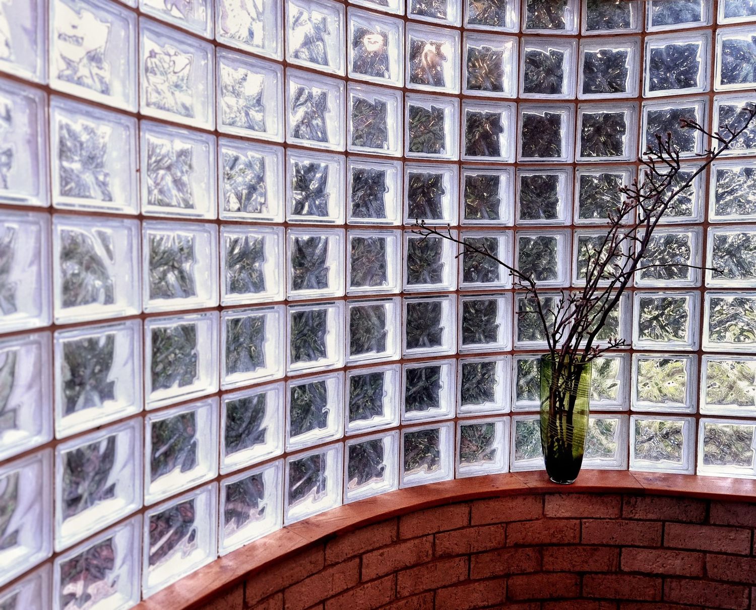 Curved glass block wall above a red brick ledge, with bare branches in a green glass vase in the corner.