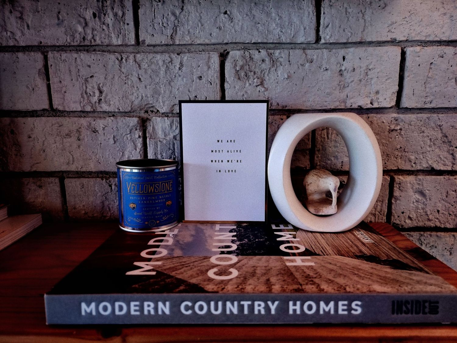 Modern Country Homes coffee table book styled with a blue candle tin, framed print and white ceramic decor against a white brick wall.