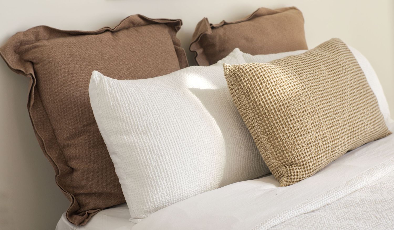 Close-up of cozy bed pillows in brown linen, white textured cotton and woven beige fabric on crisp white bedding.