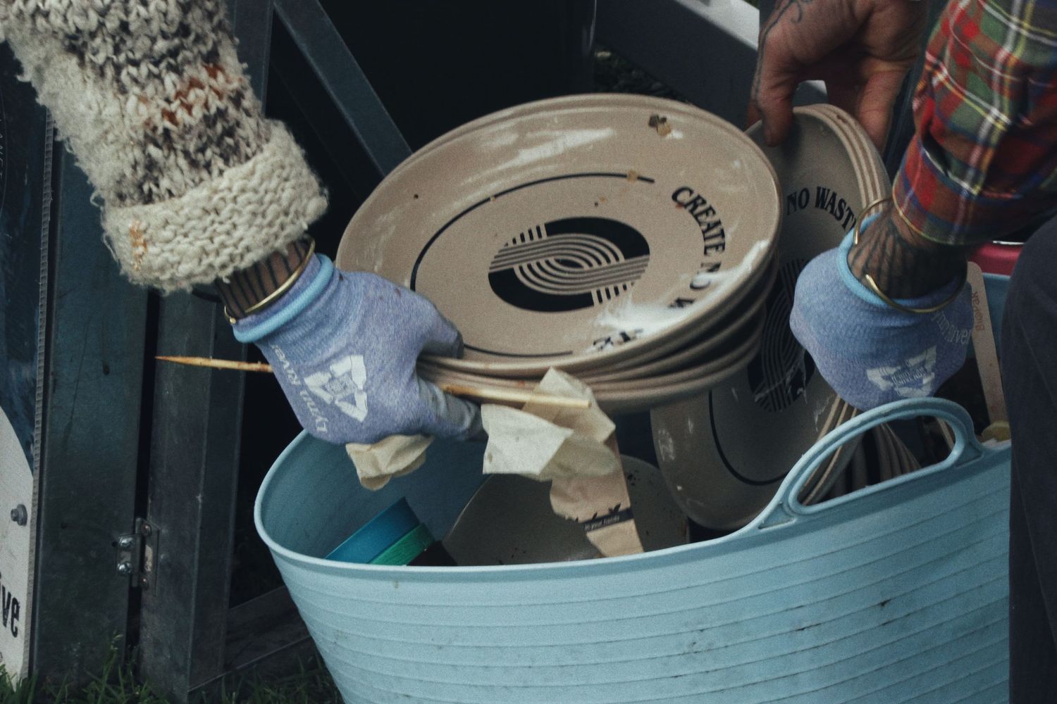 Gloved hands hold a stack of ceramic plates above a light blue plastic tub at an outdoor event.