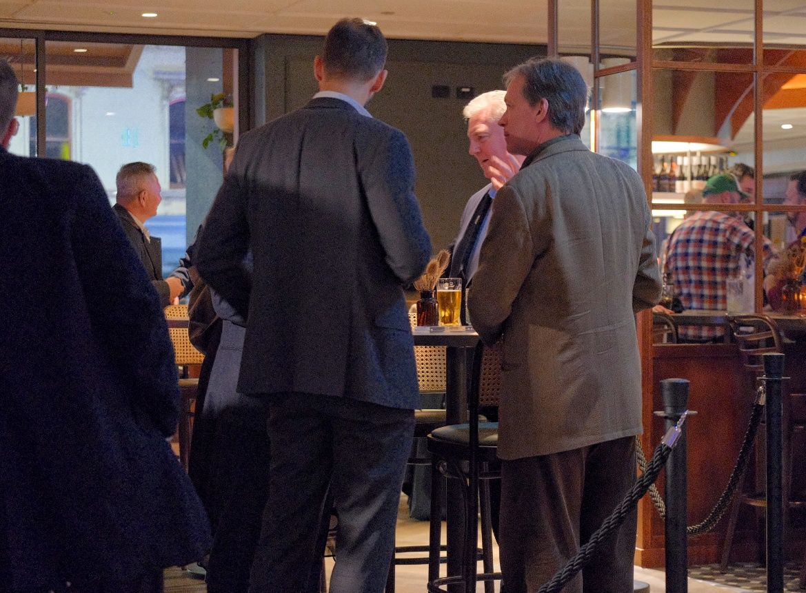 Men in business attire stand talking around a high table with drinks in a busy indoor bar or restaurant.