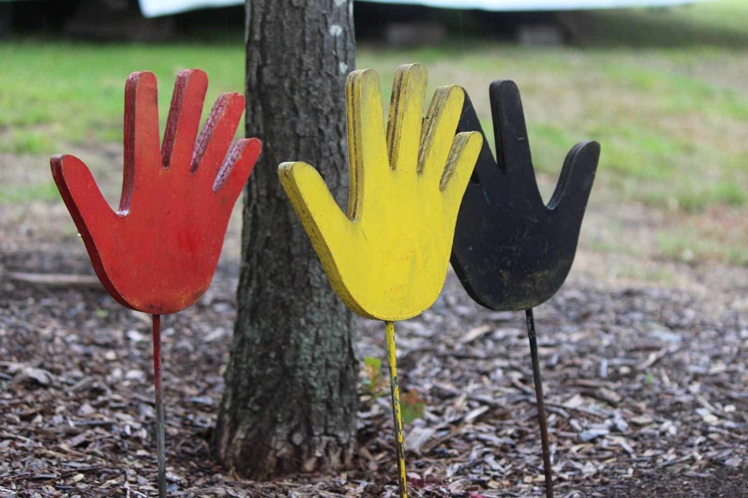 Three hand-shaped garden stakes in red, yellow and black stand on mulch beside a tree trunk with grass in the background.