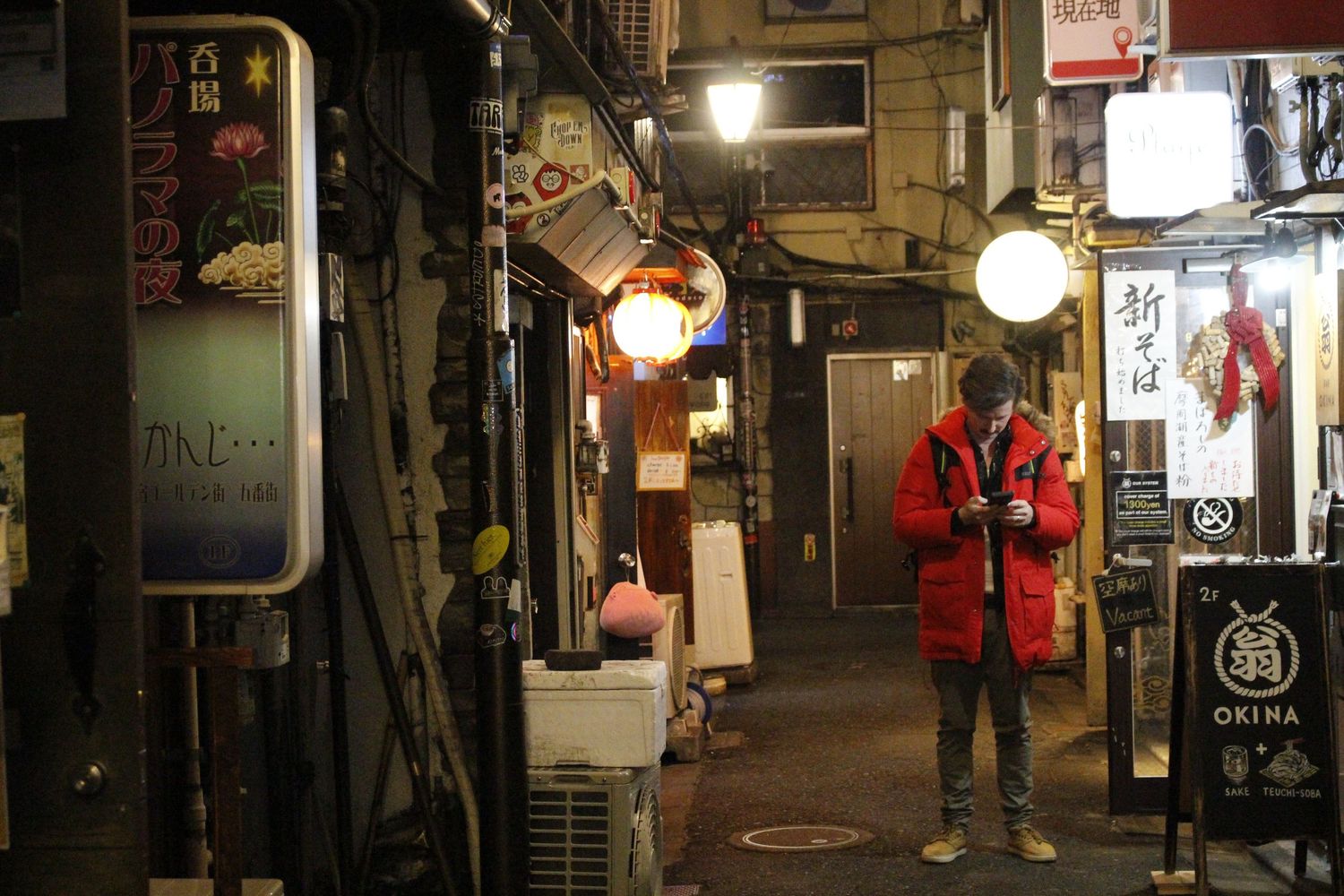 Man in a red jacket checking his phone in a narrow, lantern-lit Tokyo alley lined with small izakaya signs at night.