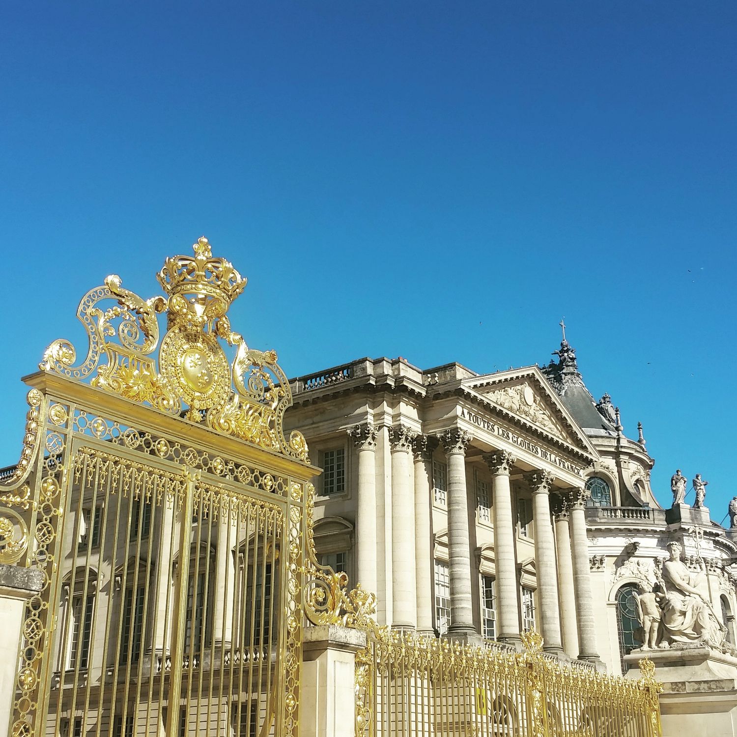 Ornate golden gates with a royal crest stand before the grand columned facade of the Palace of Versailles under a blue sky.