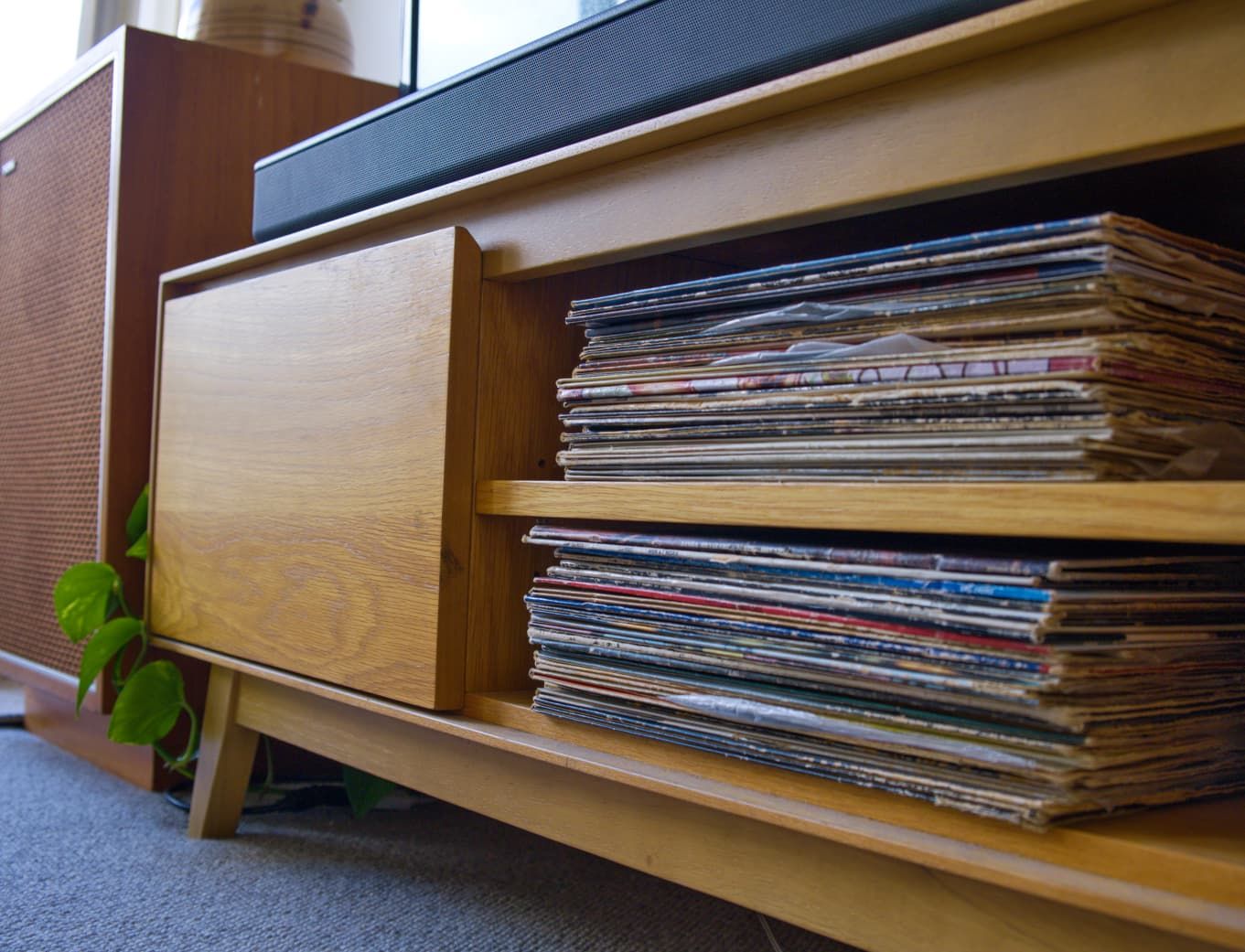 Stacks of vinyl records on a timber media console shelf with a sliding wooden door and a green plant nearby.