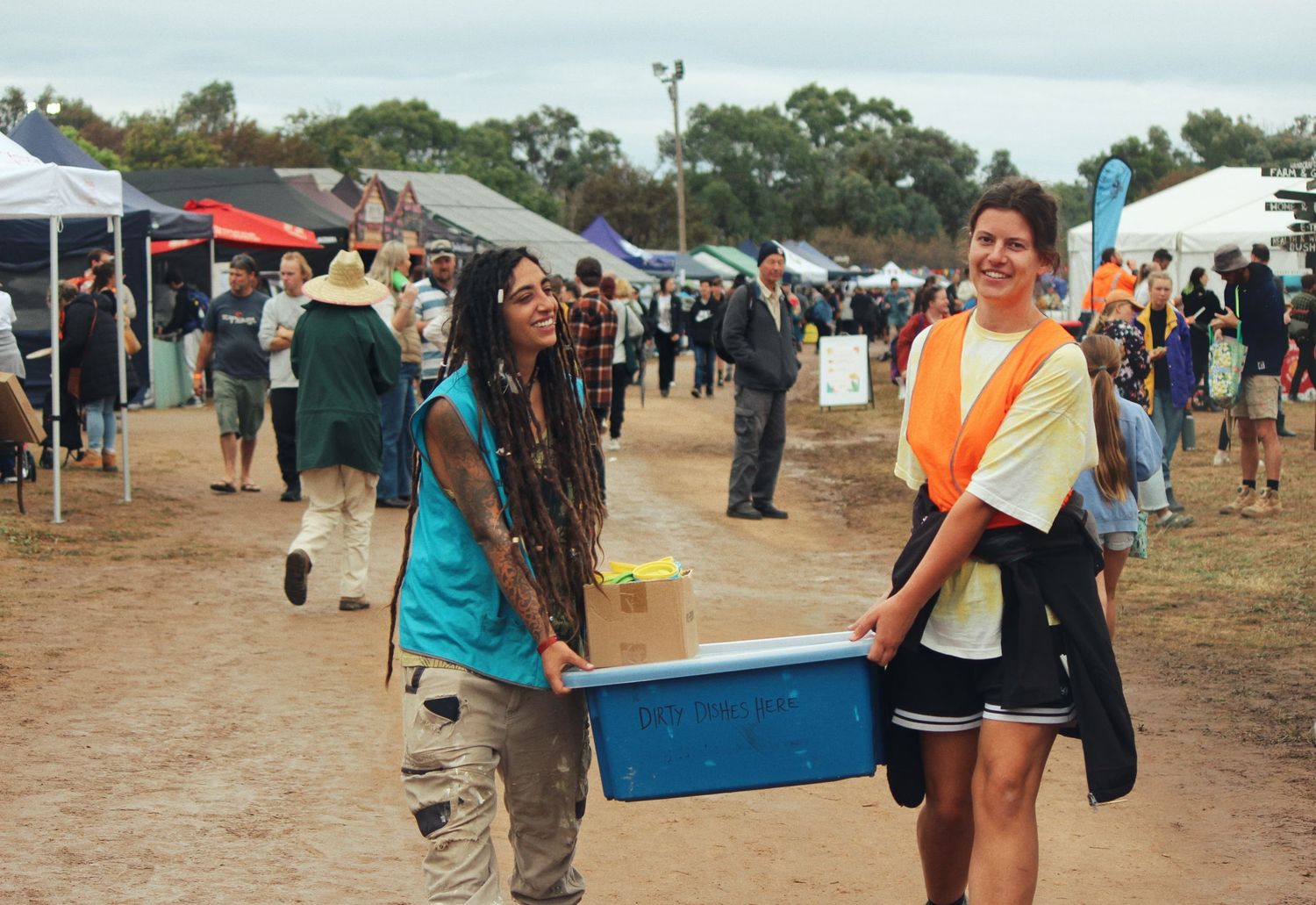Two smiling festival volunteers carry a blue plastic crate across a muddy field past tents and a crowd of people.