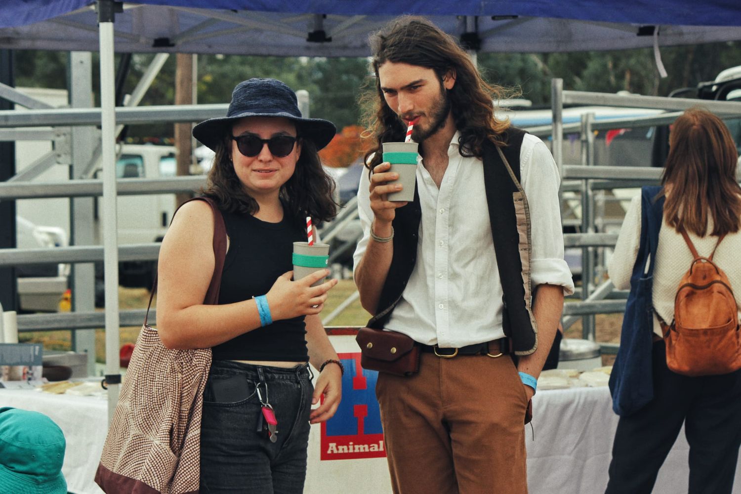 A man with long hair and a woman in a wide-brimmed hat hold drinks while standing together at an outdoor festival.