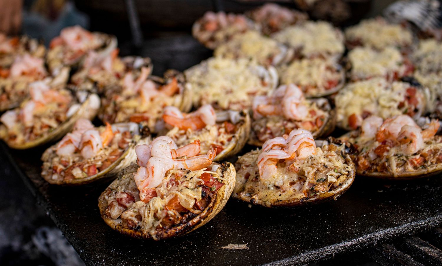 Freshly grilled seafood-topped shells with shrimp, cheese, and vegetables served on a dark cooking surface.