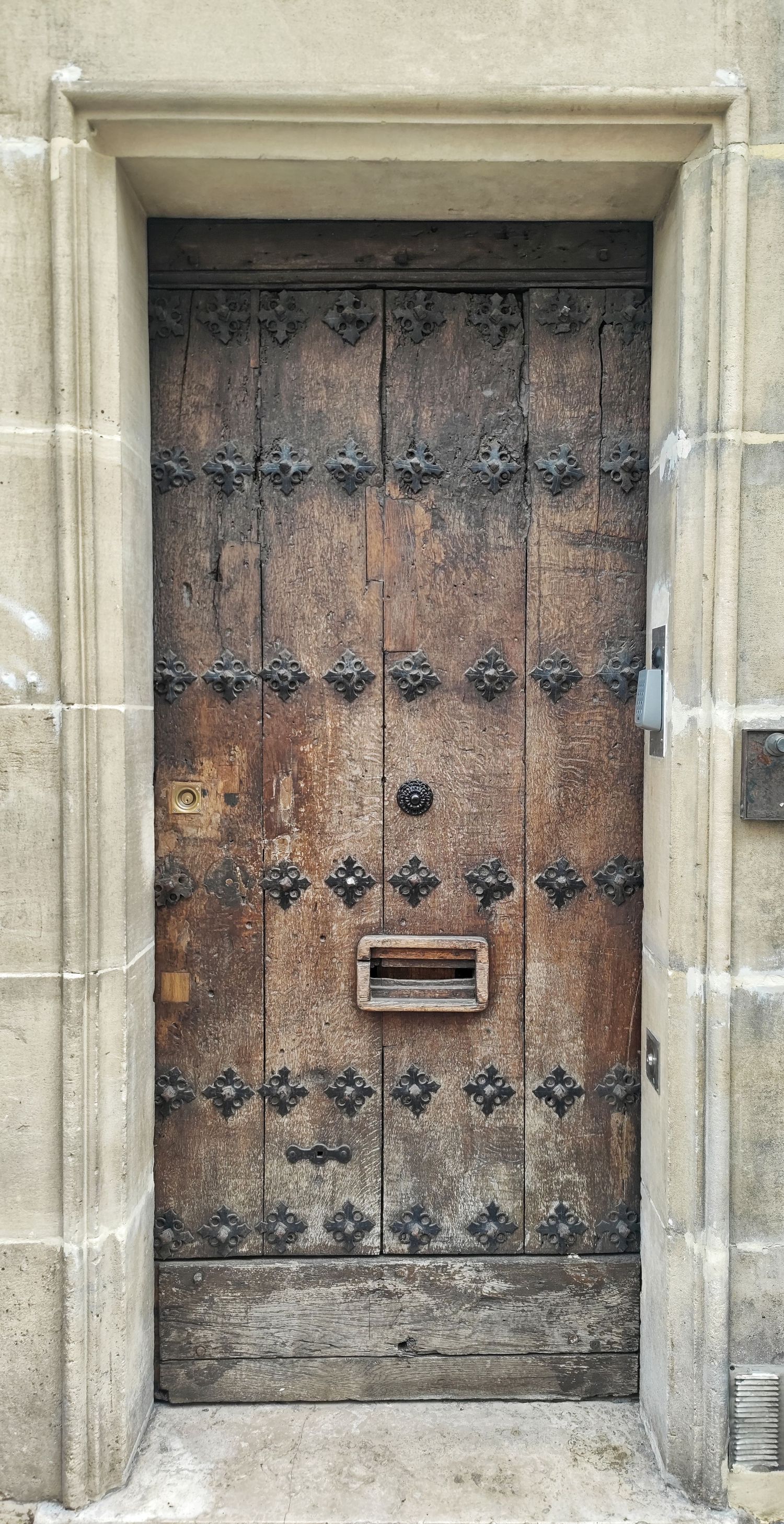Aged wooden door studded with black iron nails and a brass mail slot, set within a rustic pale stone doorway.