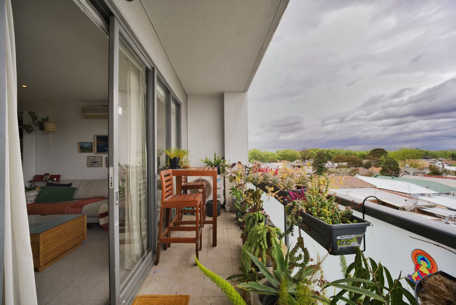 Apartment balcony with a wooden table and chairs, flowering potted plants and a view of the suburban skyline under cloudy skies.