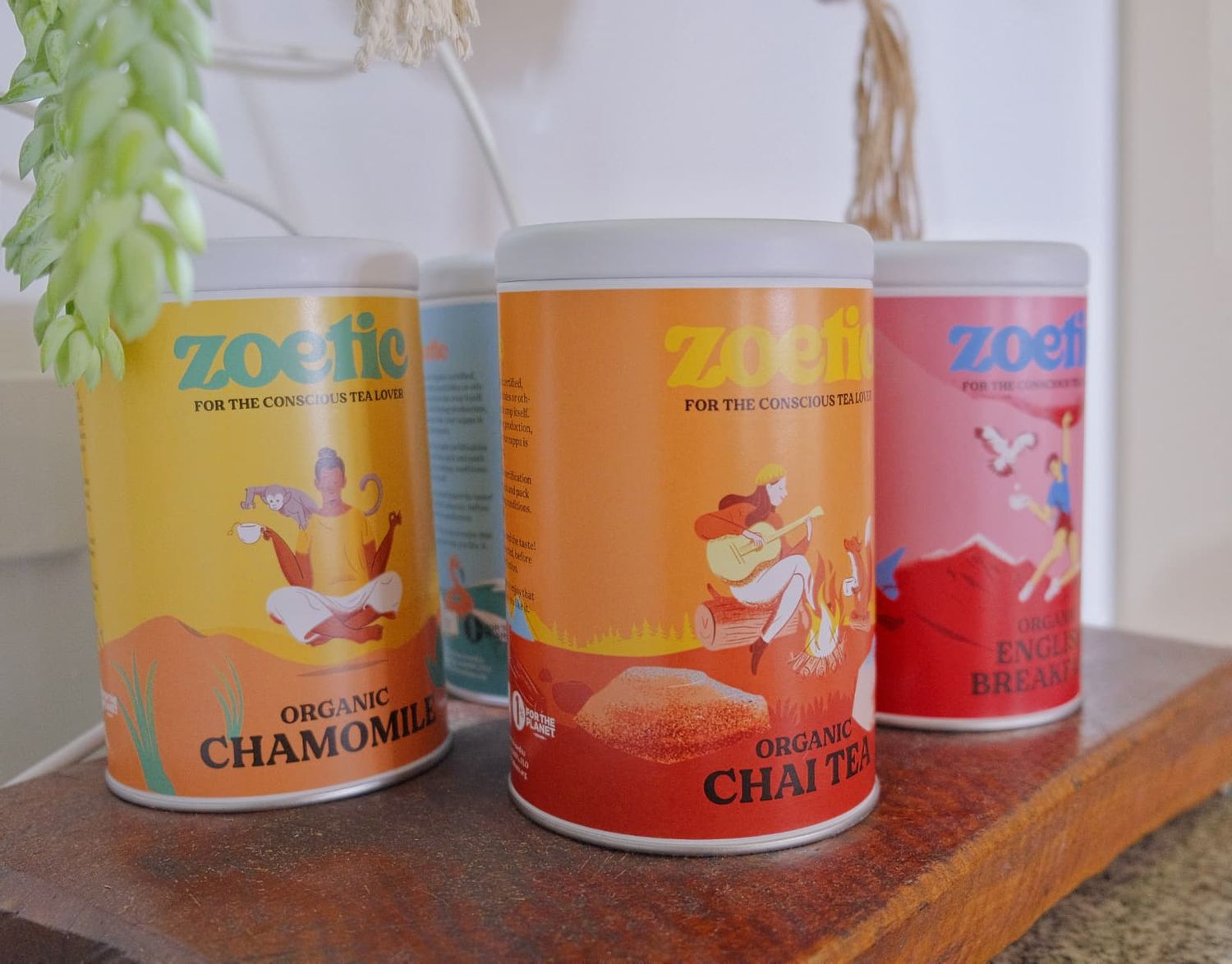 Three Zoetic organic tea tins in chamomile, chai tea and breakfast flavours with colorful illustrated labels displayed on a wooden shelf.