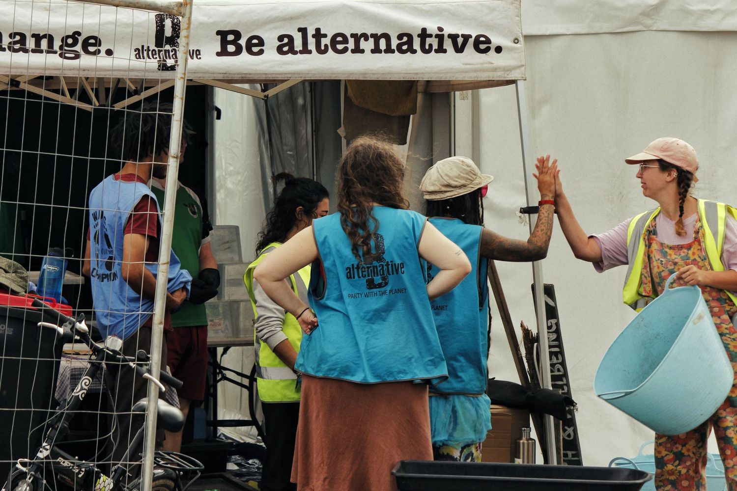Volunteers in blue and yellow vests chat beside a festival tent marked with the slogan Be alternative, holding a blue tub.