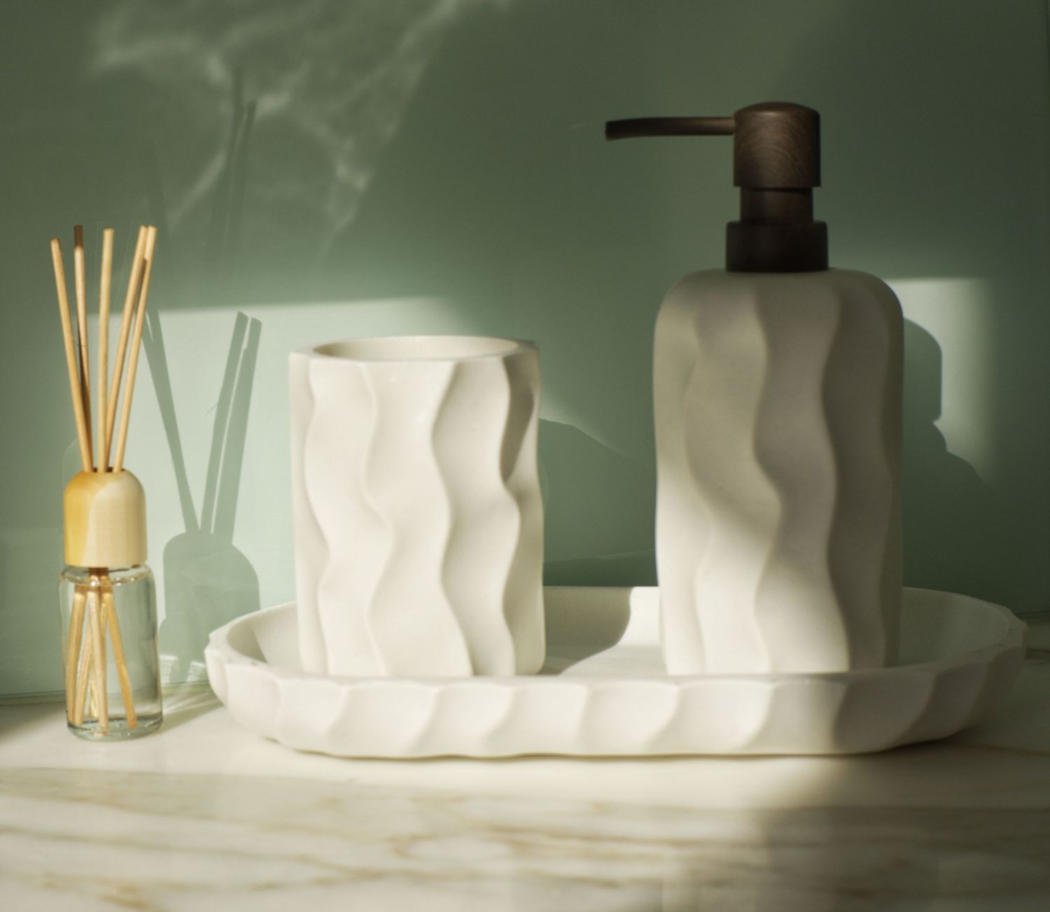 White wavy-textured bathroom set with soap dispenser, tumbler and tray beside a reed diffuser on a marble countertop.