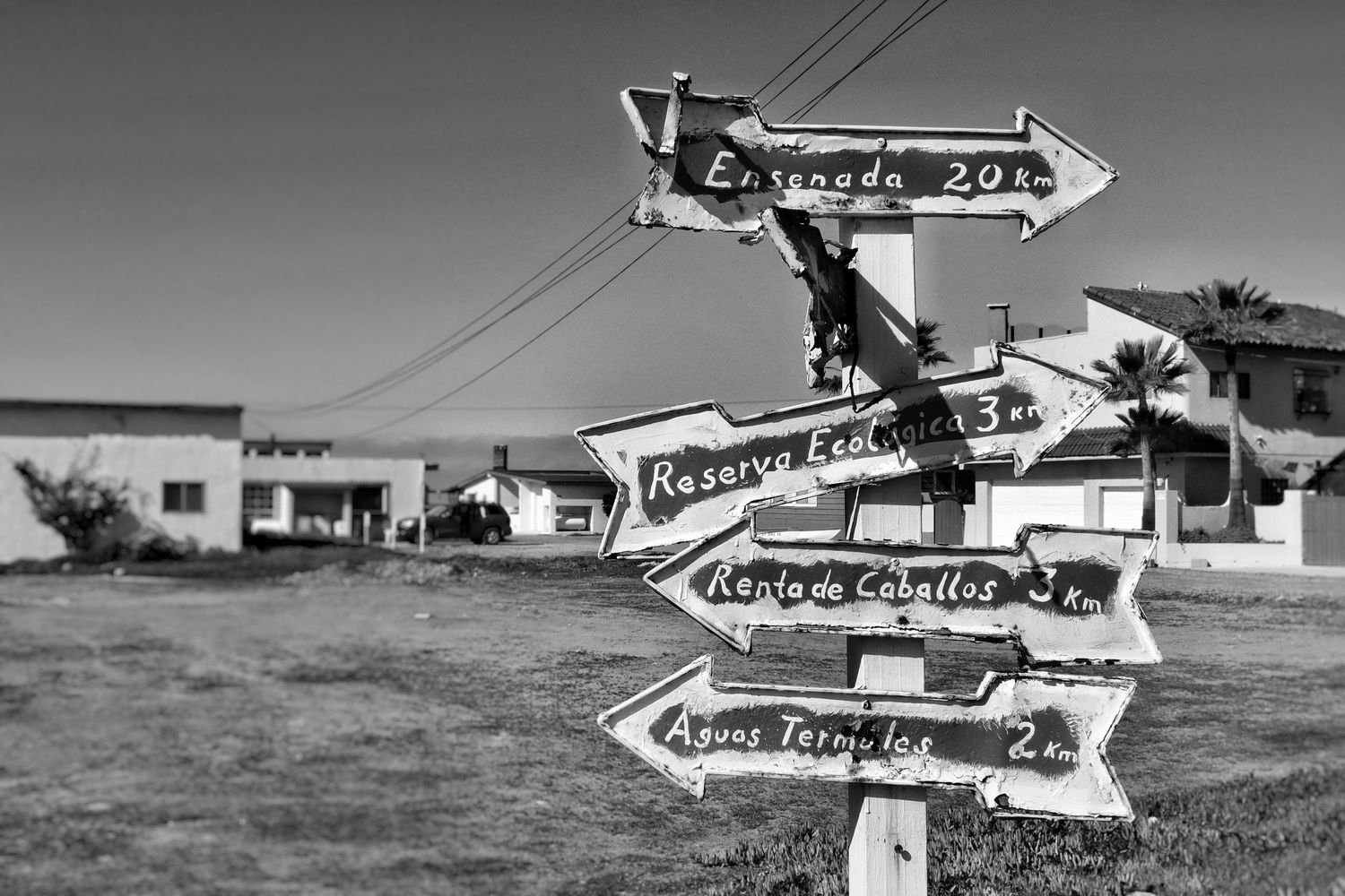 Black and white photo of a wooden signpost with arrows pointing to Ensenada, horseback rentals, and hot springs on a dusty coastal road.