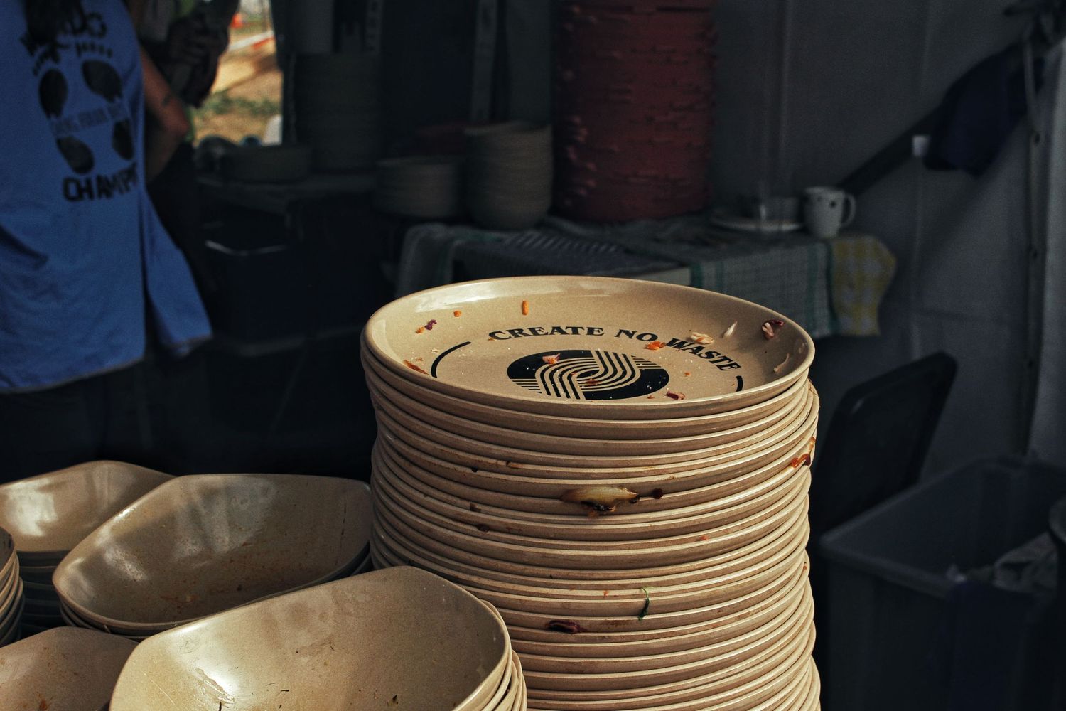 Stack of reusable wooden-style plates stamped with a Create No Waste logo sits on a festival food stall counter.