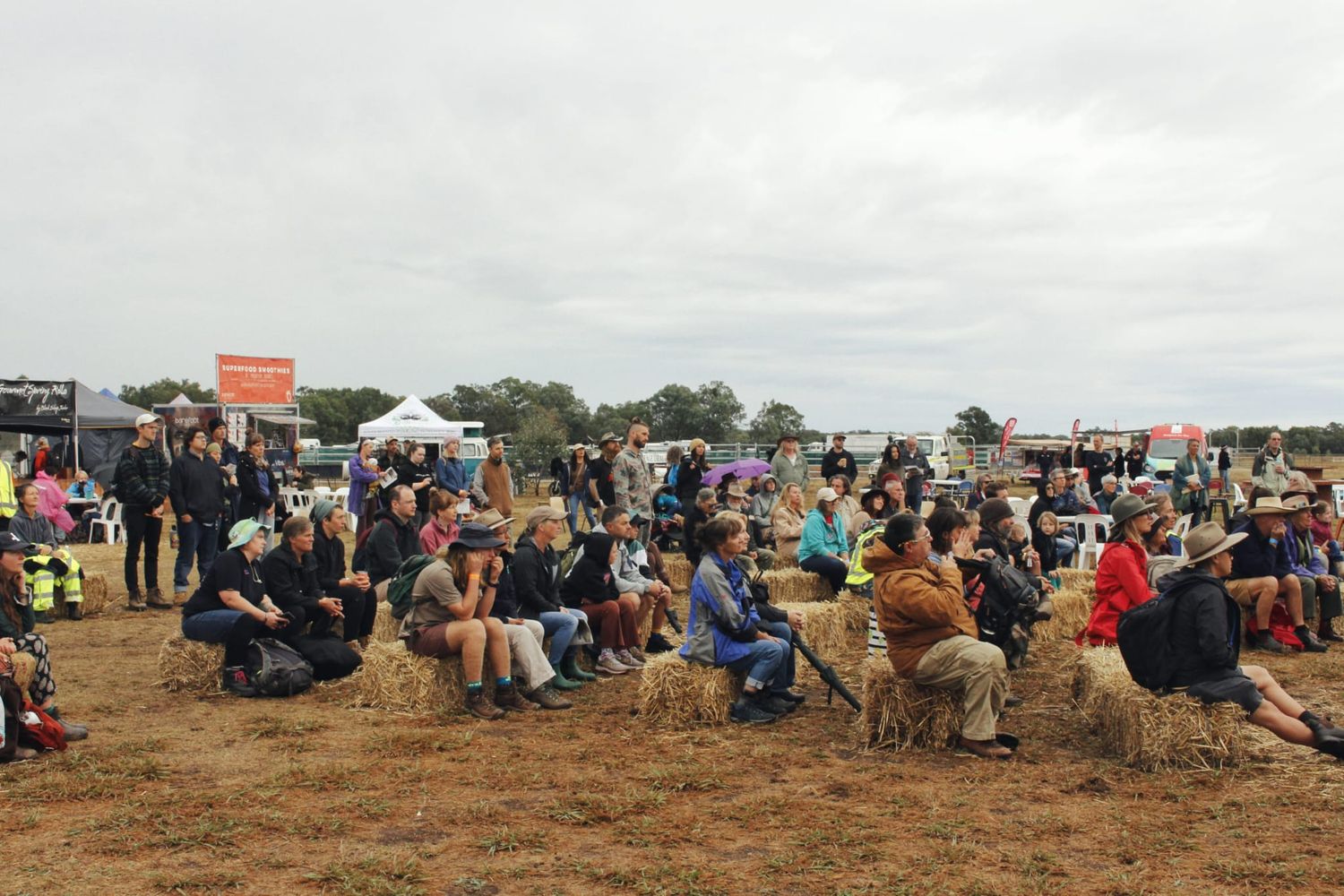 A seated crowd gathers on hay bales in a dry field to watch a talk or performance at an outdoor festival under an overcast sky.