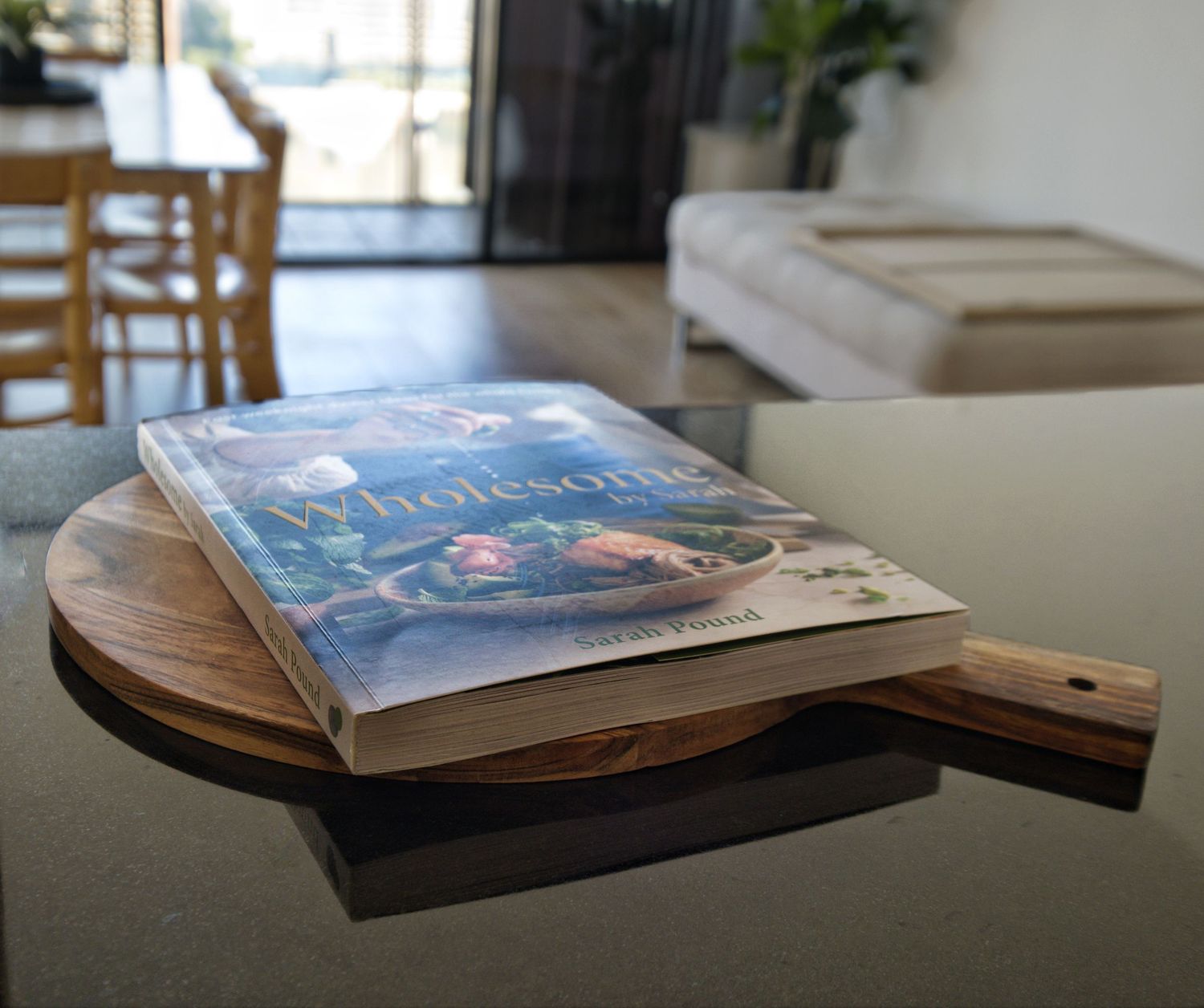 Cookbook resting on a round wooden serving board on a glossy black benchtop with the dining area in the background.