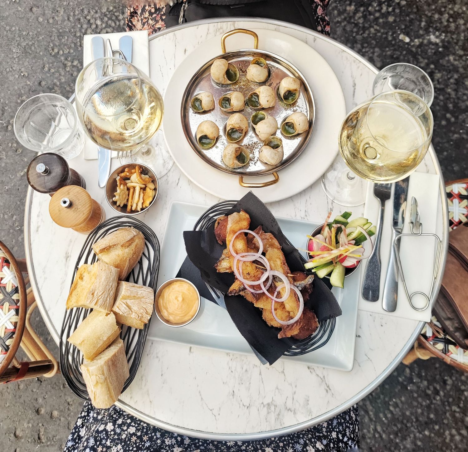 Overhead view of a Parisian cafe table with escargots, sliced baguette, sausage with onions, salad, and two glasses of white wine.
