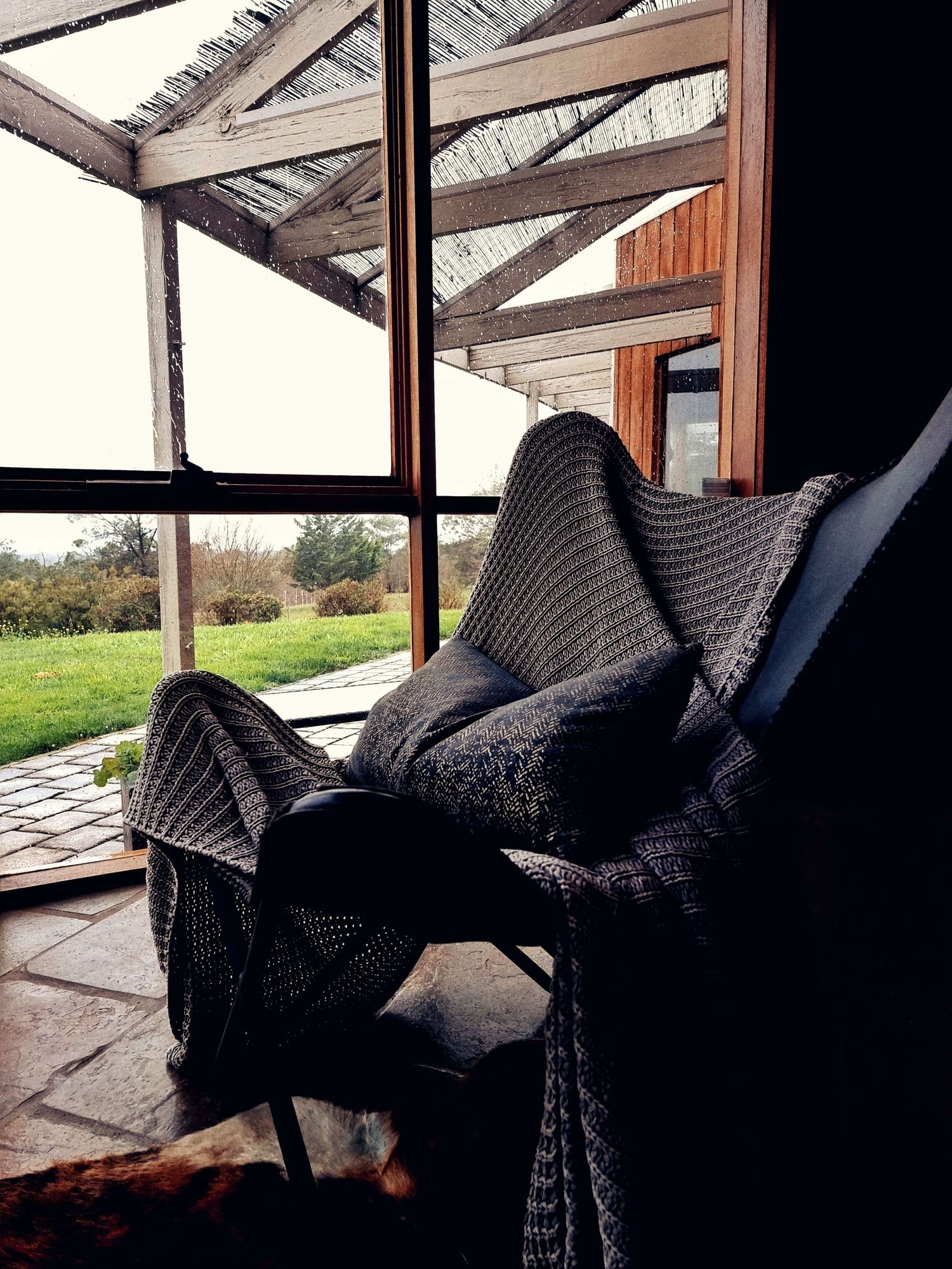 Cozy knitted blanket and cushion on a chair beside a large window overlooking a green lawn and a pergola with timber beams.