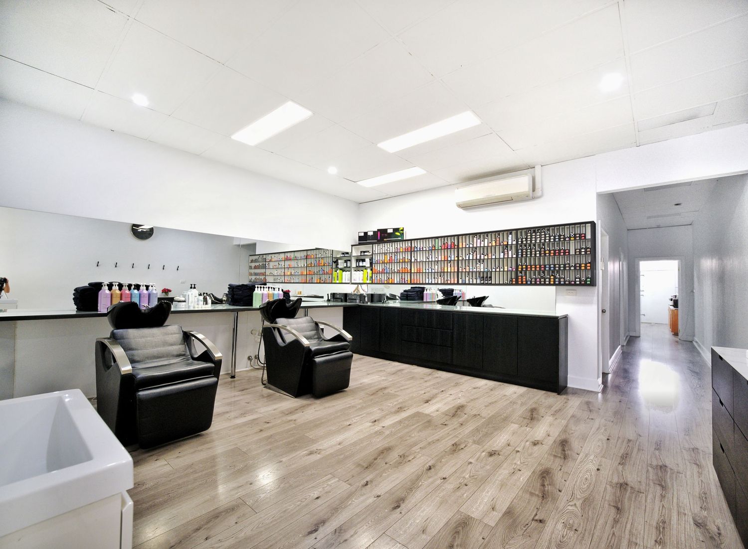 Long hair salon interior with a wall of hair color swatches, black shampoo chairs, a dark reception counter and wood-look flooring.