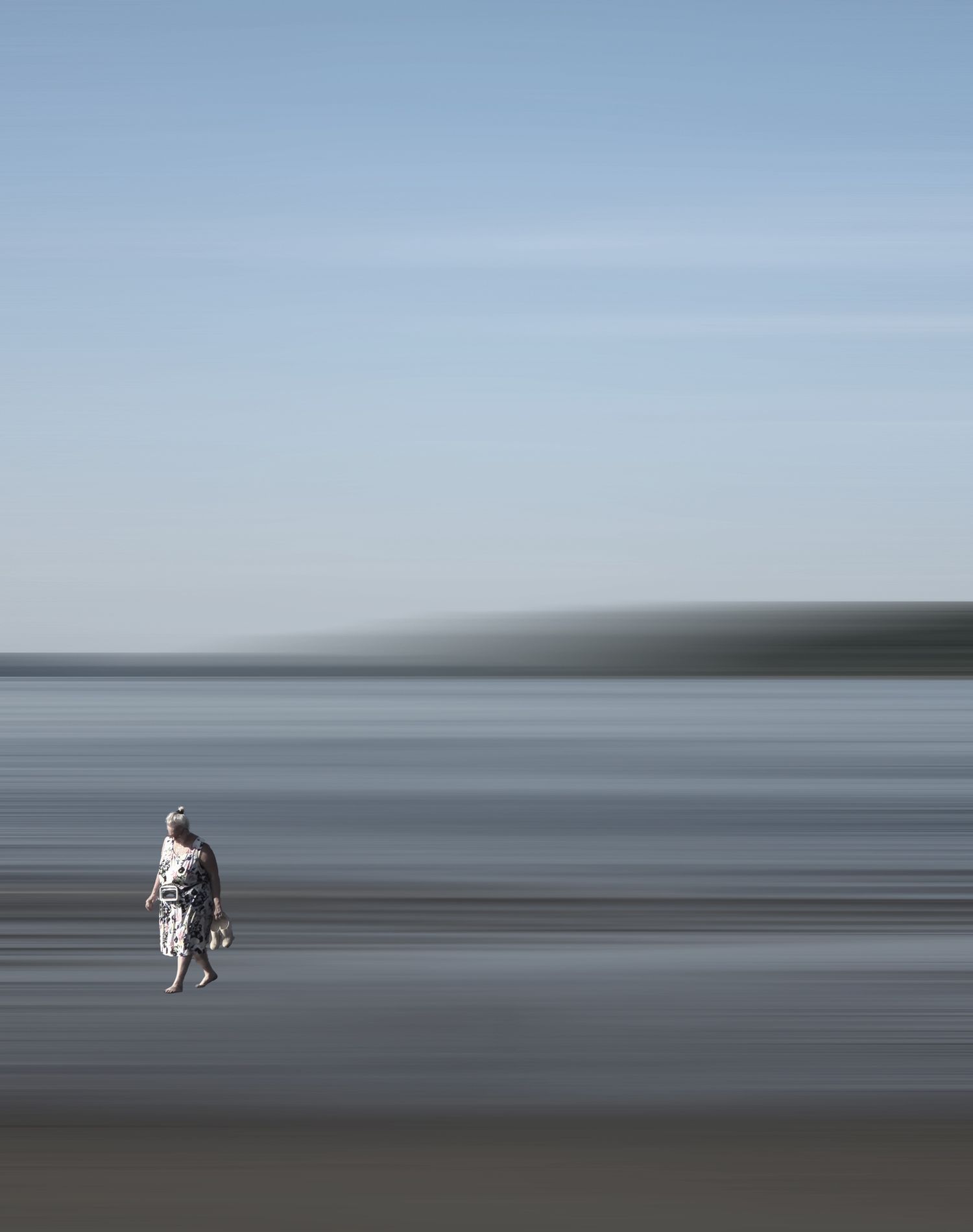 A lone figure in a patterned dress walks along a blurred beach under a pale blue sky.