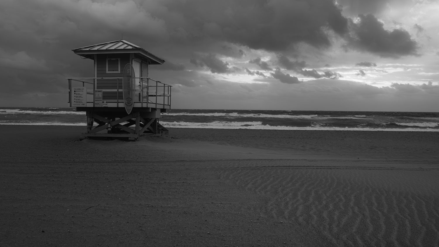 Black and white beach scene with a lifeguard tower on empty sand under dramatic stormy skies.