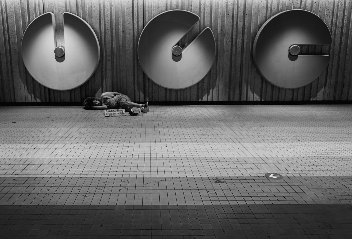 Black and white photo of a homeless person sleeping on a subway platform beneath an art sculpture.