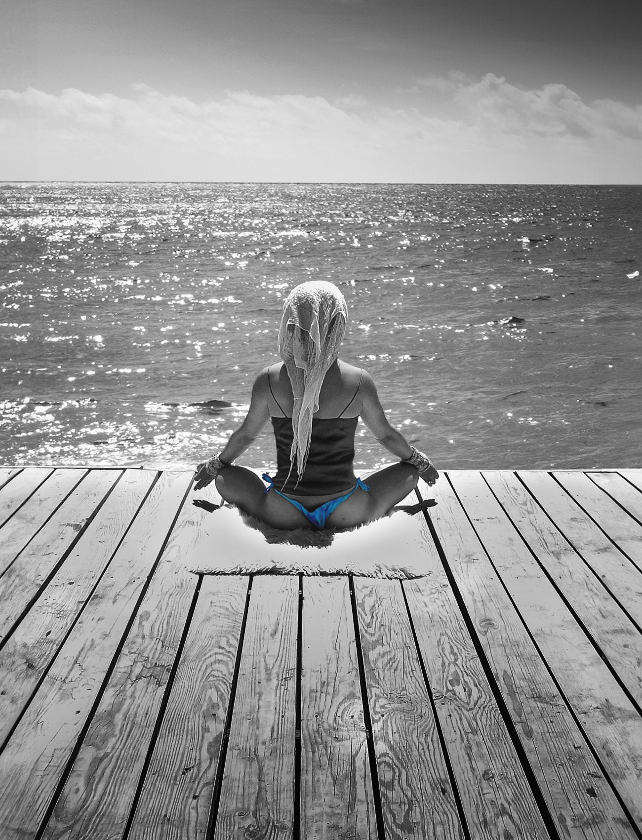 Black and white selective color image of a blonde woman in blue bikini meditating on a seaside dock.
