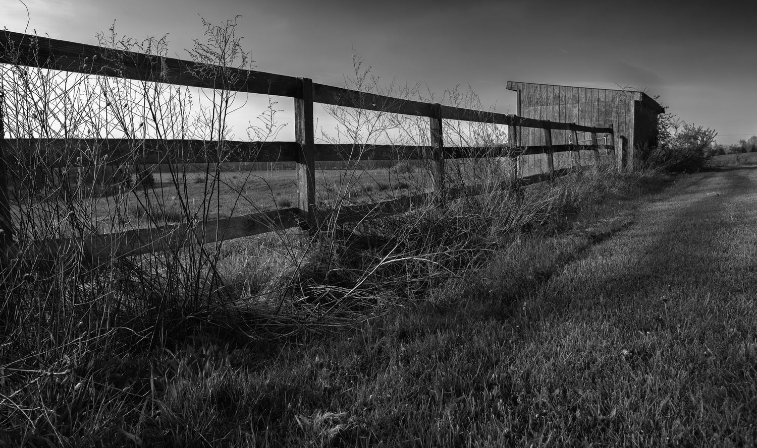 Black and white rural landscape featuring a wooden fence leading to an old barn in open fields.