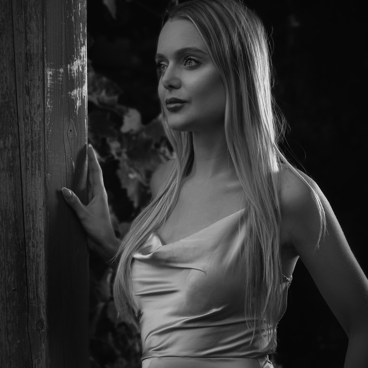 Elegant black and white portrait of a young blonde woman in satin top leaning against wooden post.