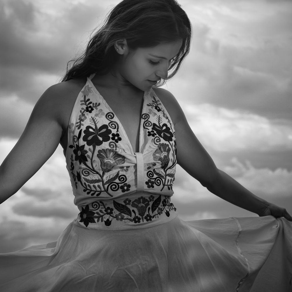 Young woman in an embroidered halter dress twirling outdoors under a dramatic cloudy sky.