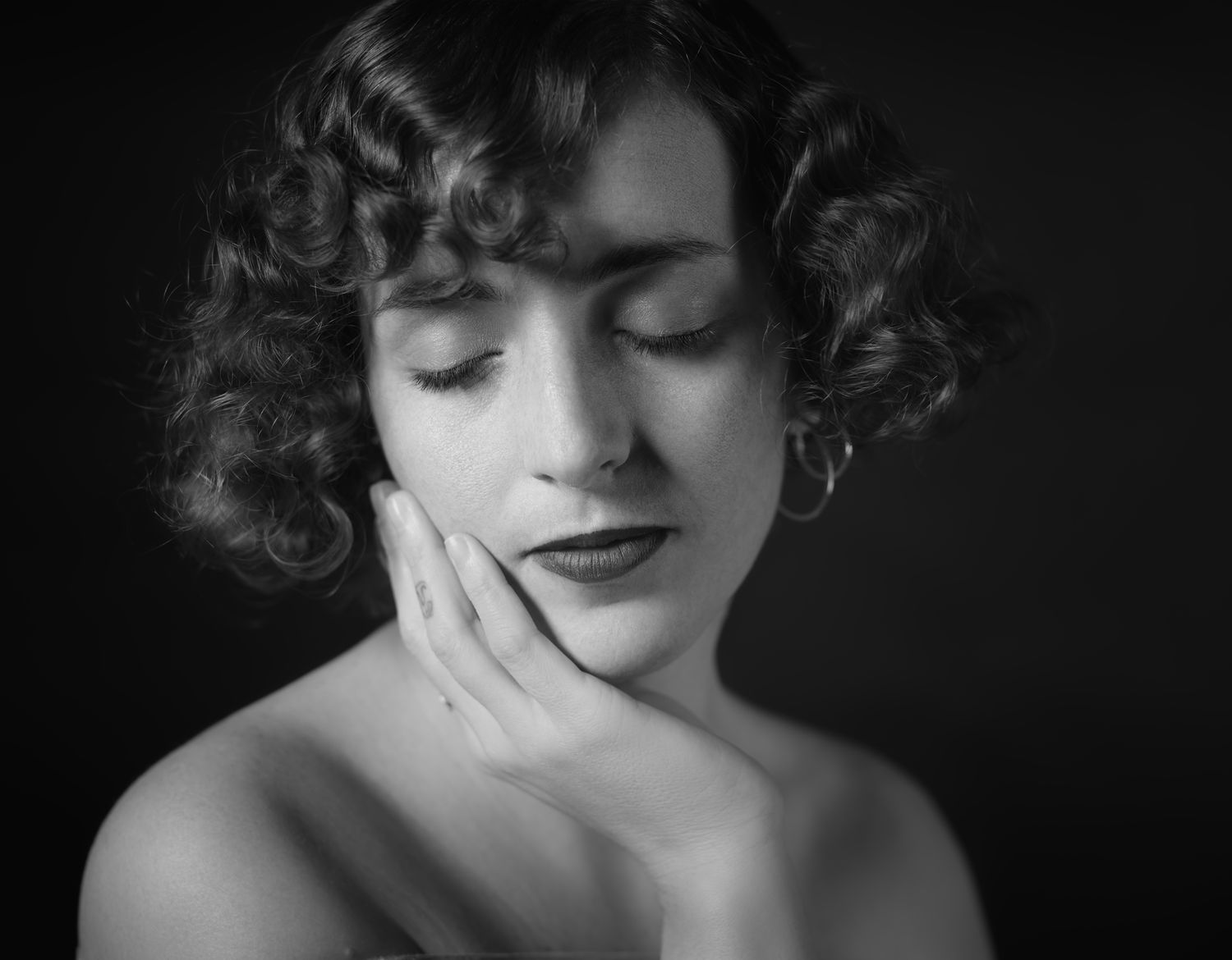 Black and white portrait of a young woman with curly hair, eyes closed, hand gently touching her cheek.