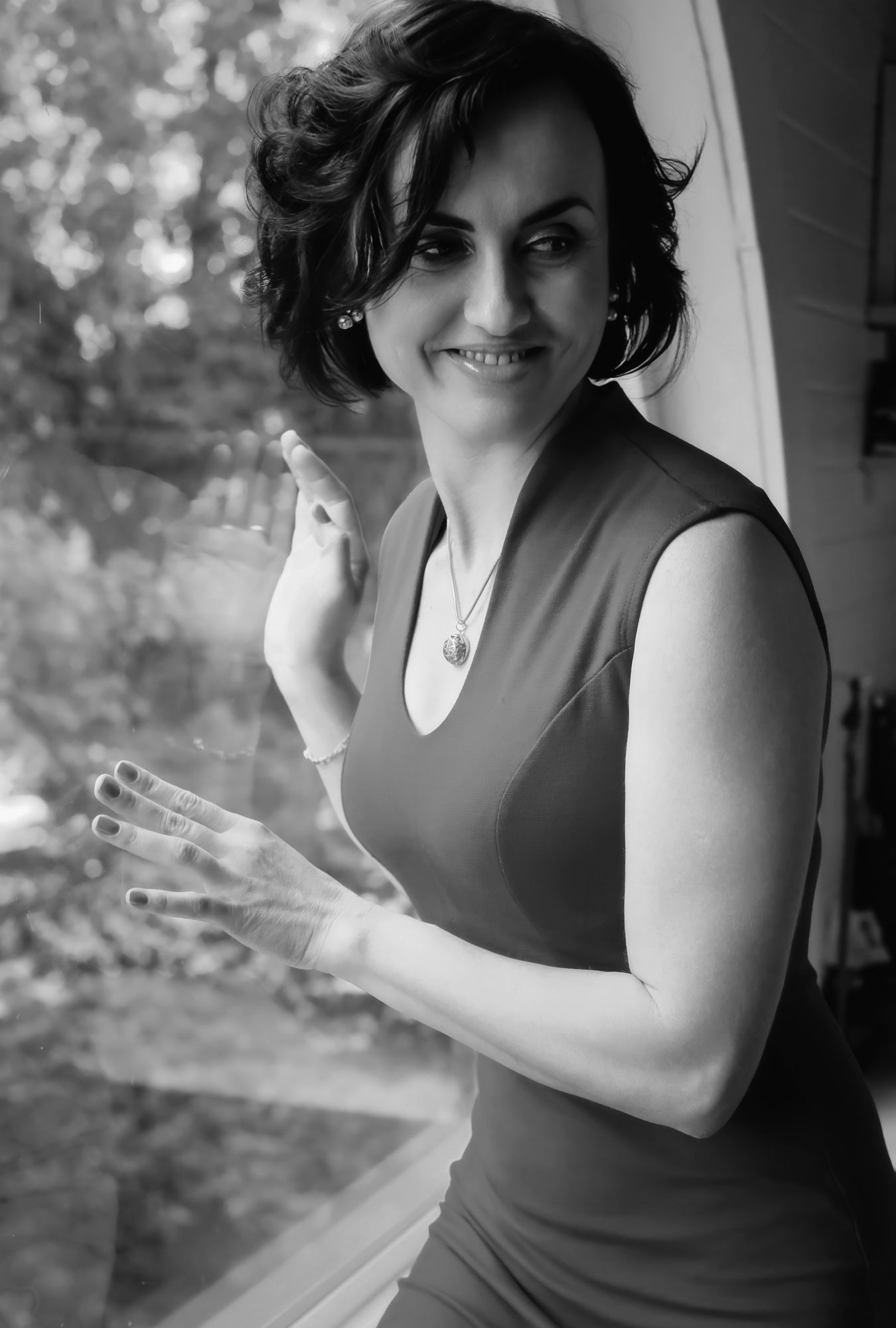 Black and white portrait of a smiling woman in sleeveless dress, posing by a window with hand raised.