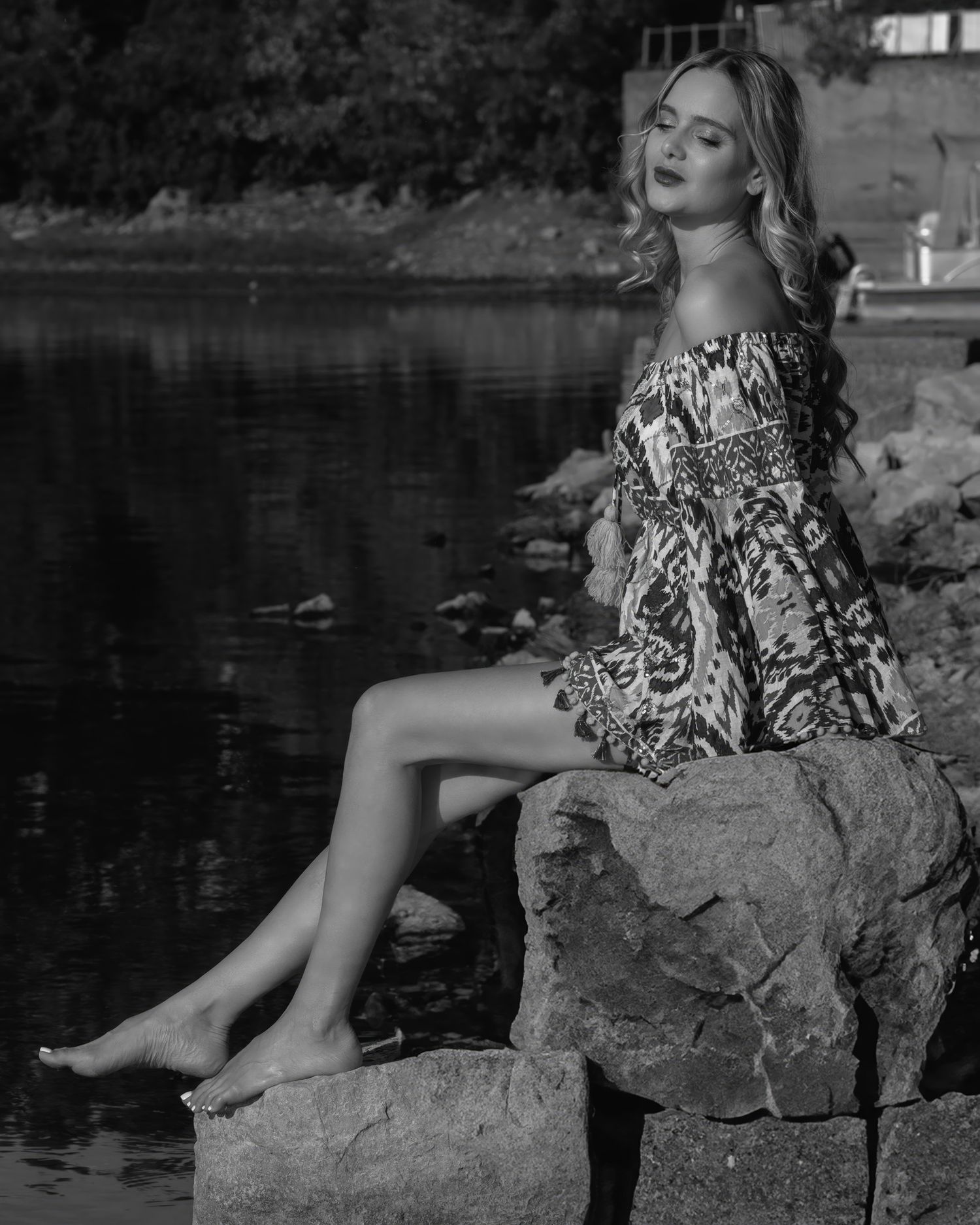 Black and white photo of a young woman in a floral off-shoulder dress sitting barefoot on rocks by water.