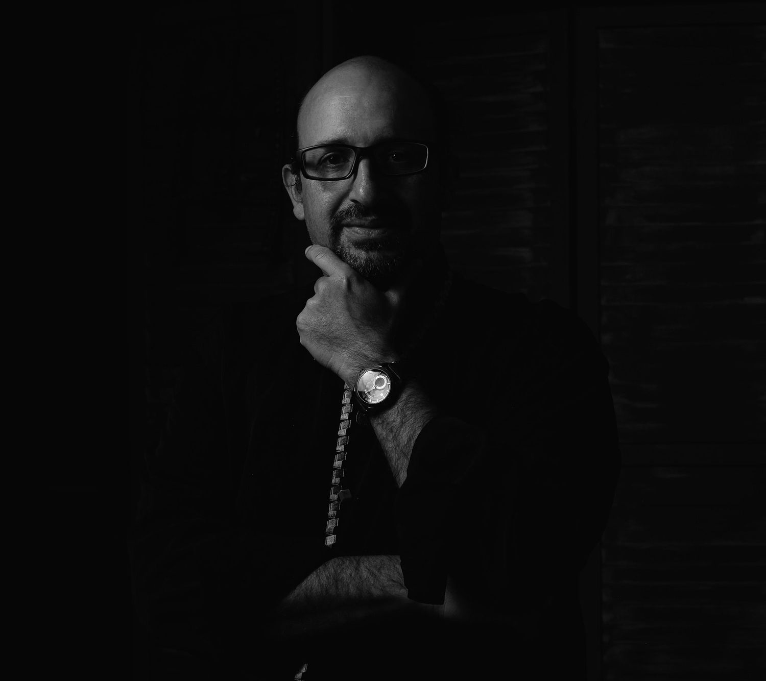 Dramatic black and white portrait of a bald man with glasses and a watch in thoughtful pose.