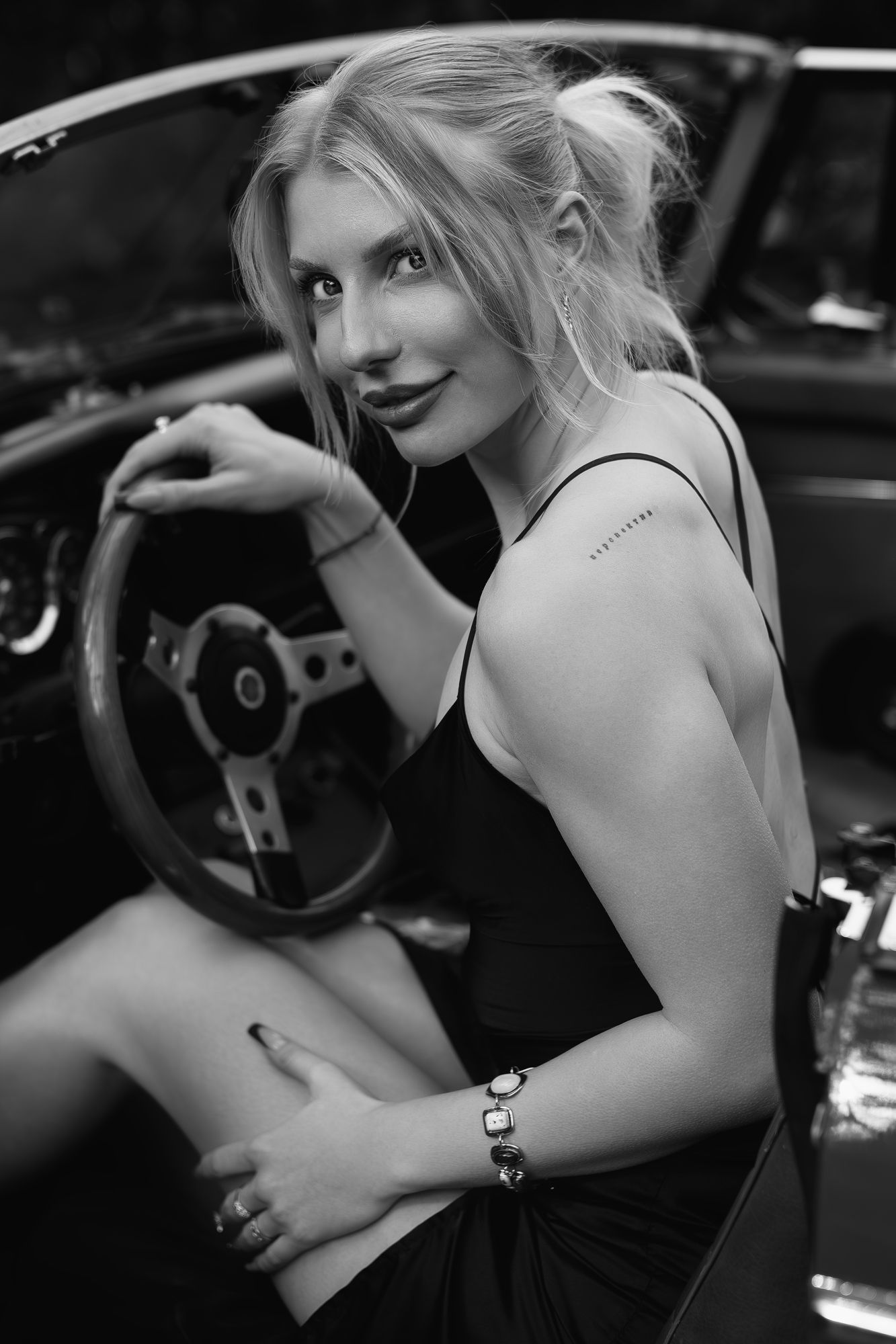 Black and white photo of a blonde woman in black dress sitting in a vintage car holding steering wheel.