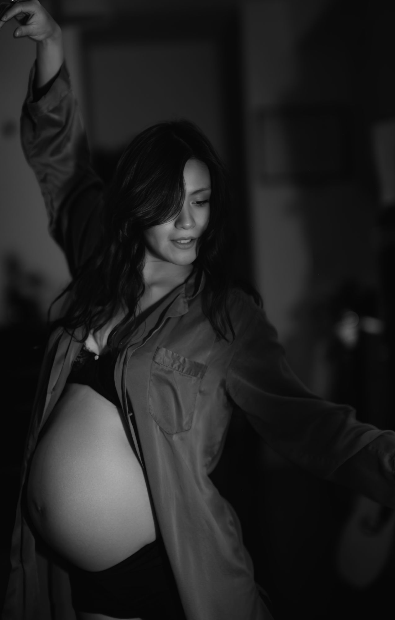 Black and white photo of a smiling pregnant woman posing with arms raised, wearing an open shirt revealing her baby bump.