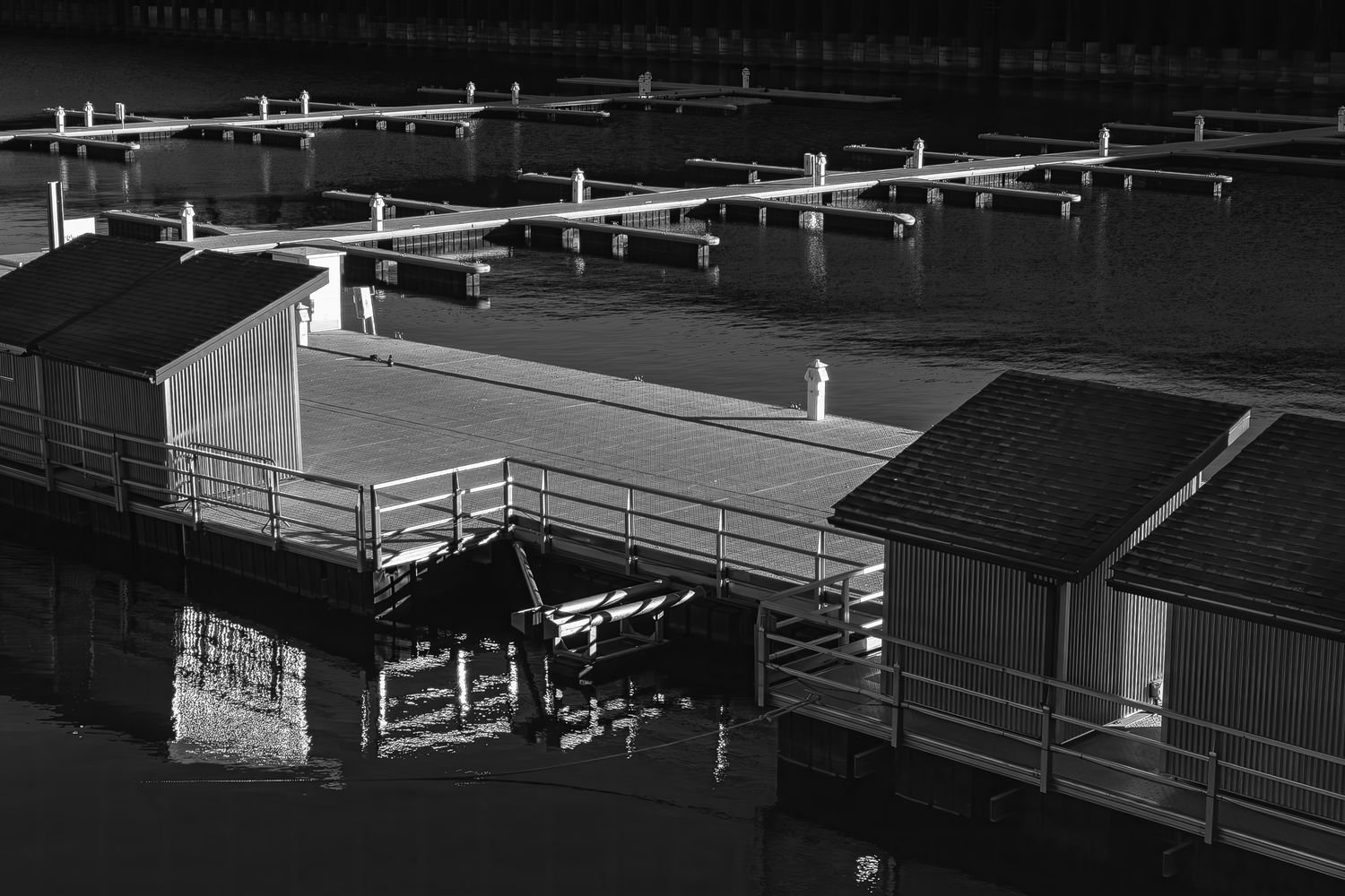 Black and white aerial view of floating boat docks and boathouses reflected in still marina water.