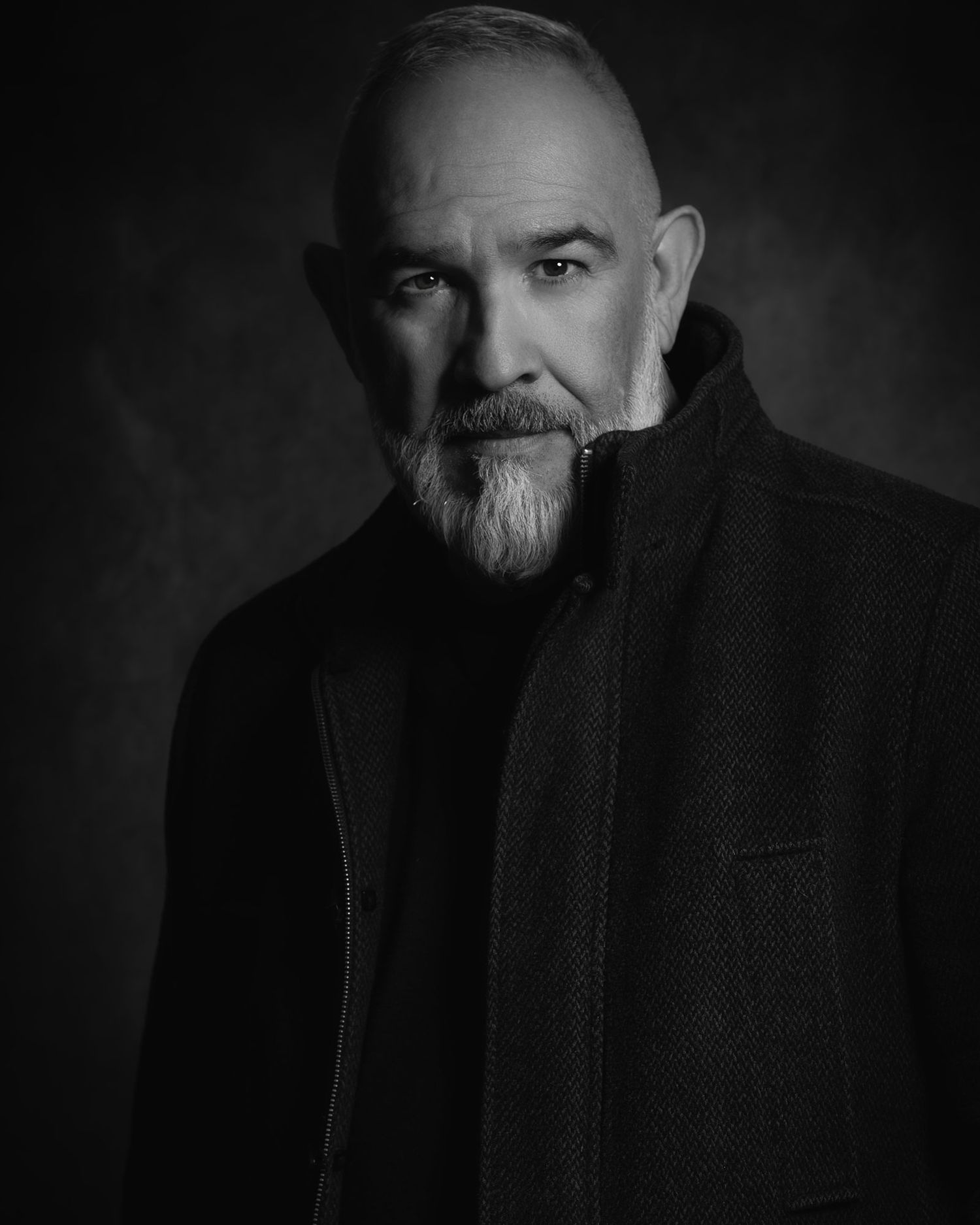 Black and white dramatic portrait of a bald older man with goatee wearing a dark coat.
