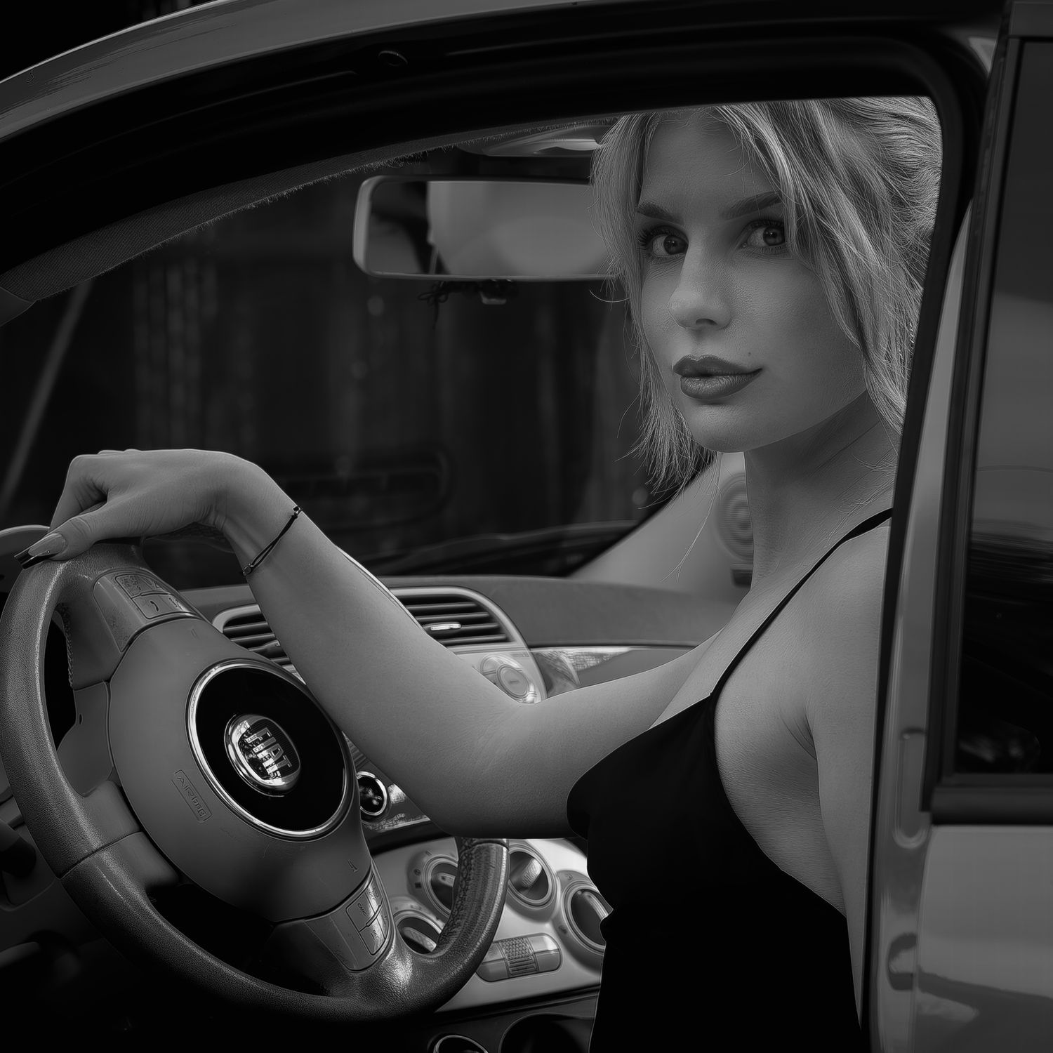 Blonde woman in a black dress sitting in a car holding the steering wheel, black and white photo.